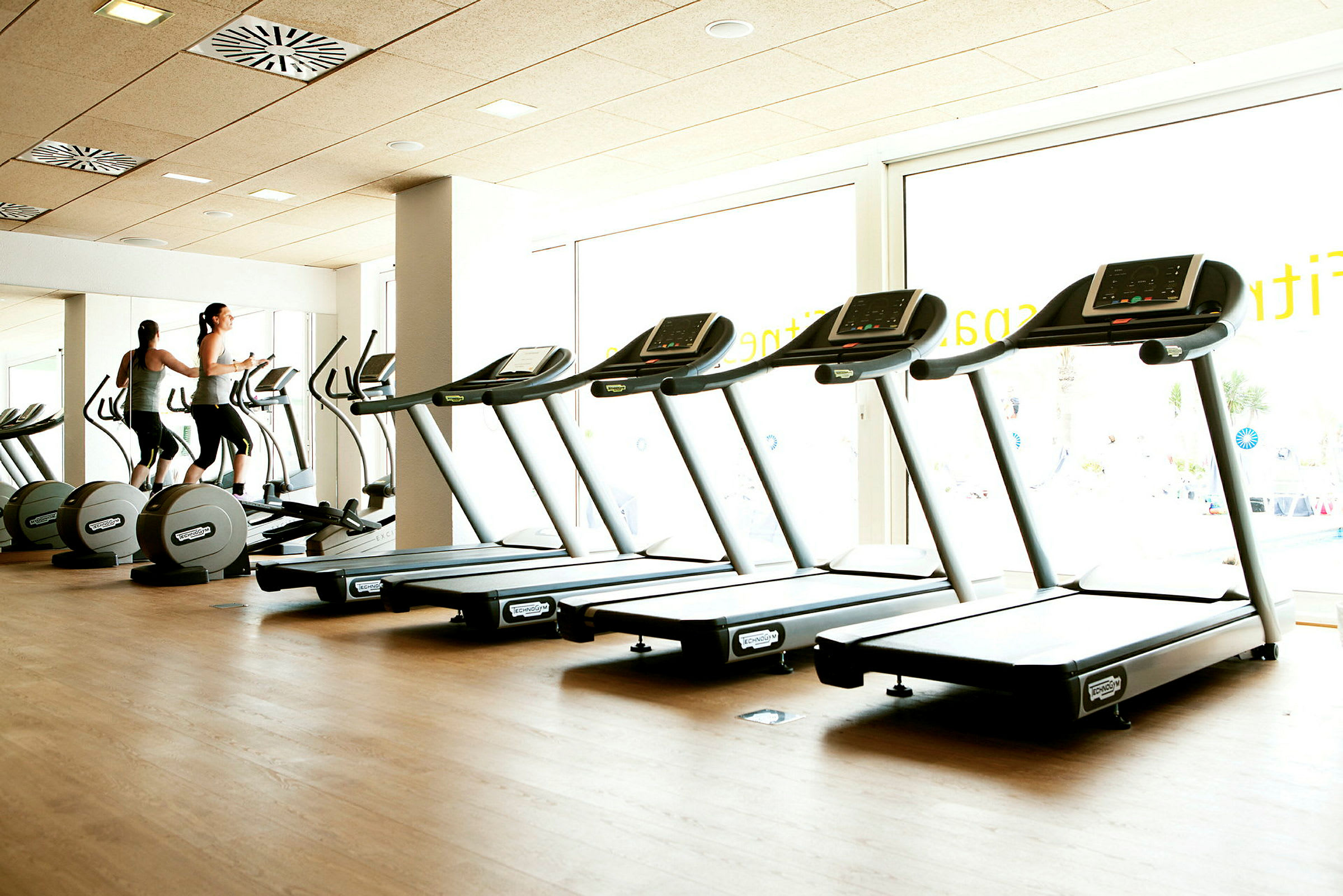 Et fitnesscenter med løbebånd og motionscykler, lys indretning med store vinduer. (AI-genereret tekst)