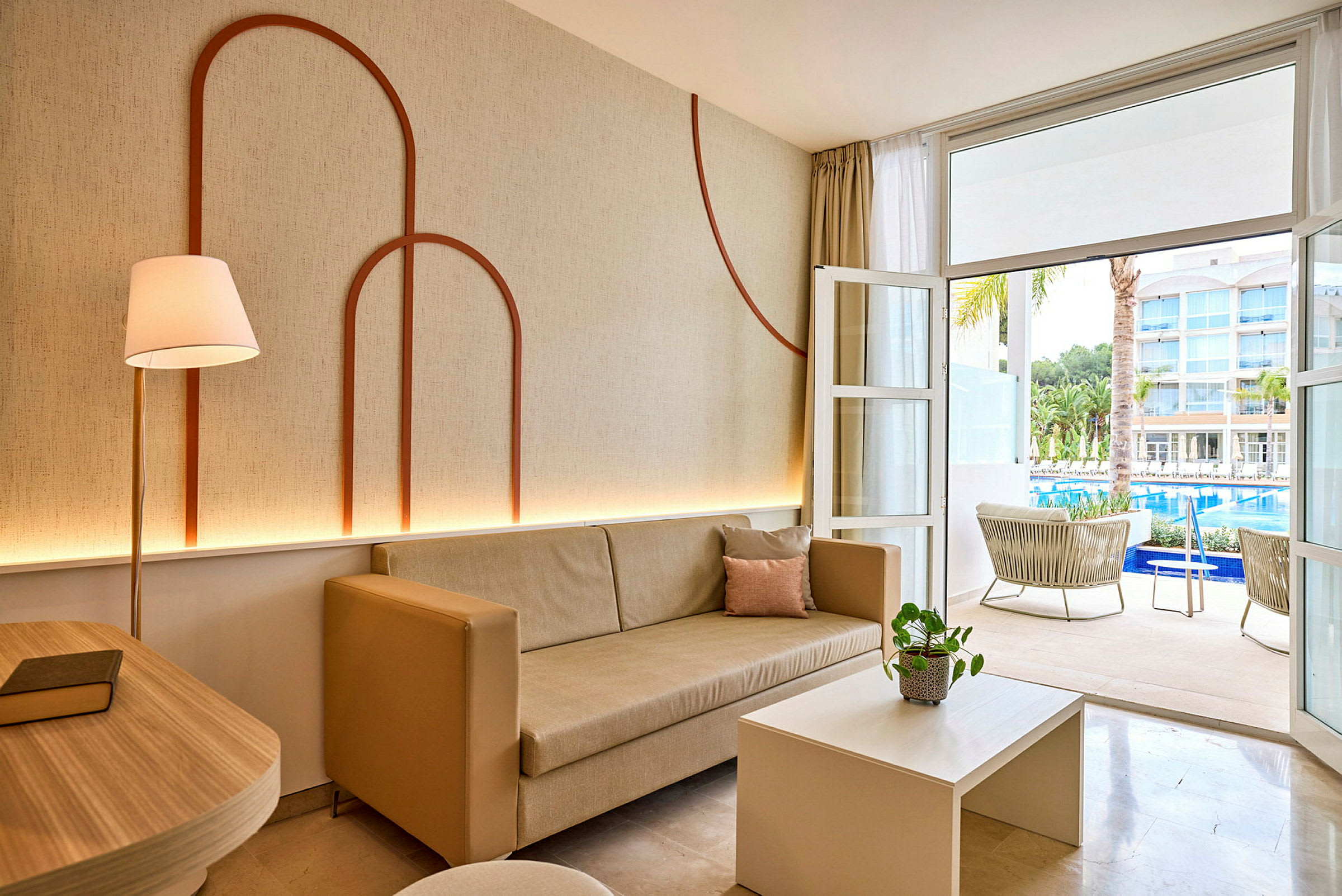 Stue med beige sofa og dekoreret væg, åbning mod terrasse med udsigt til pool og hotelbygninger. (AI-genereret tekst)