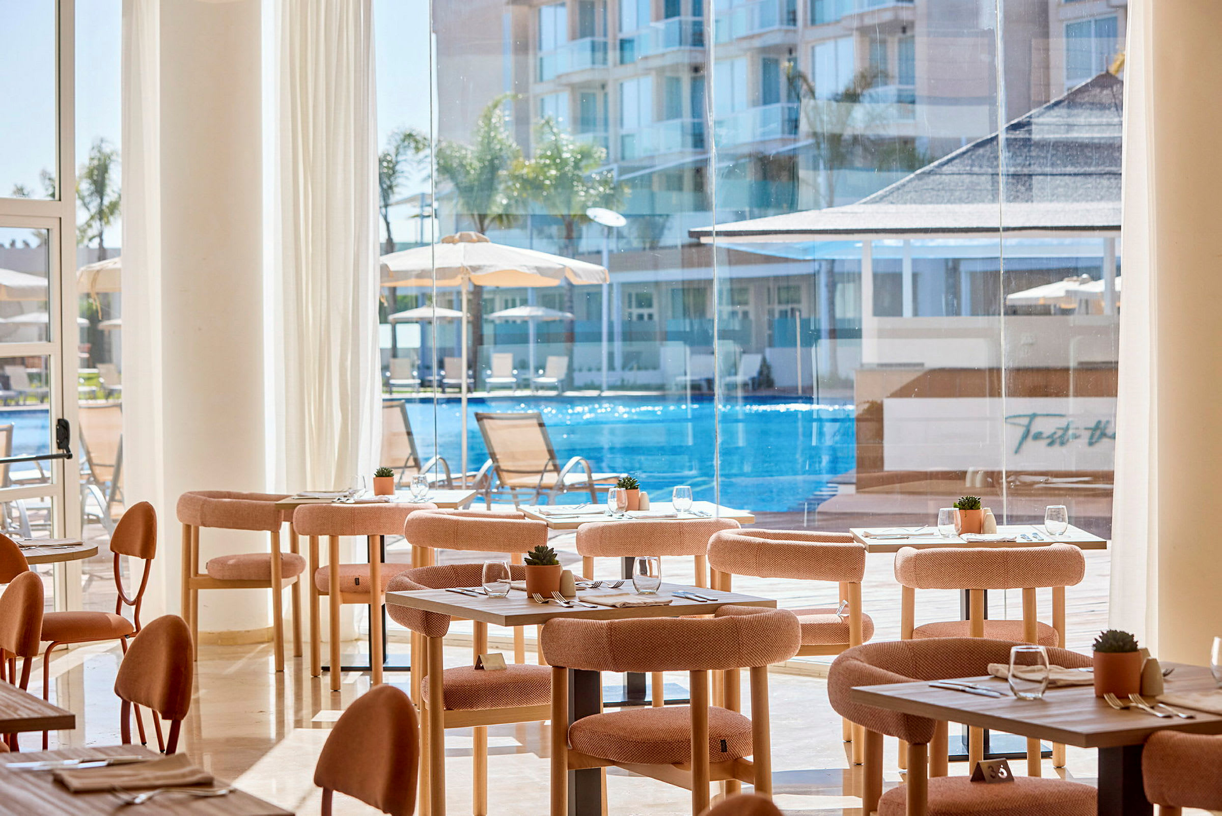 Interiør af en restaurant med borde og stole, store vinduer med udsigt over en pool og bygninger. (AI-genereret tekst)