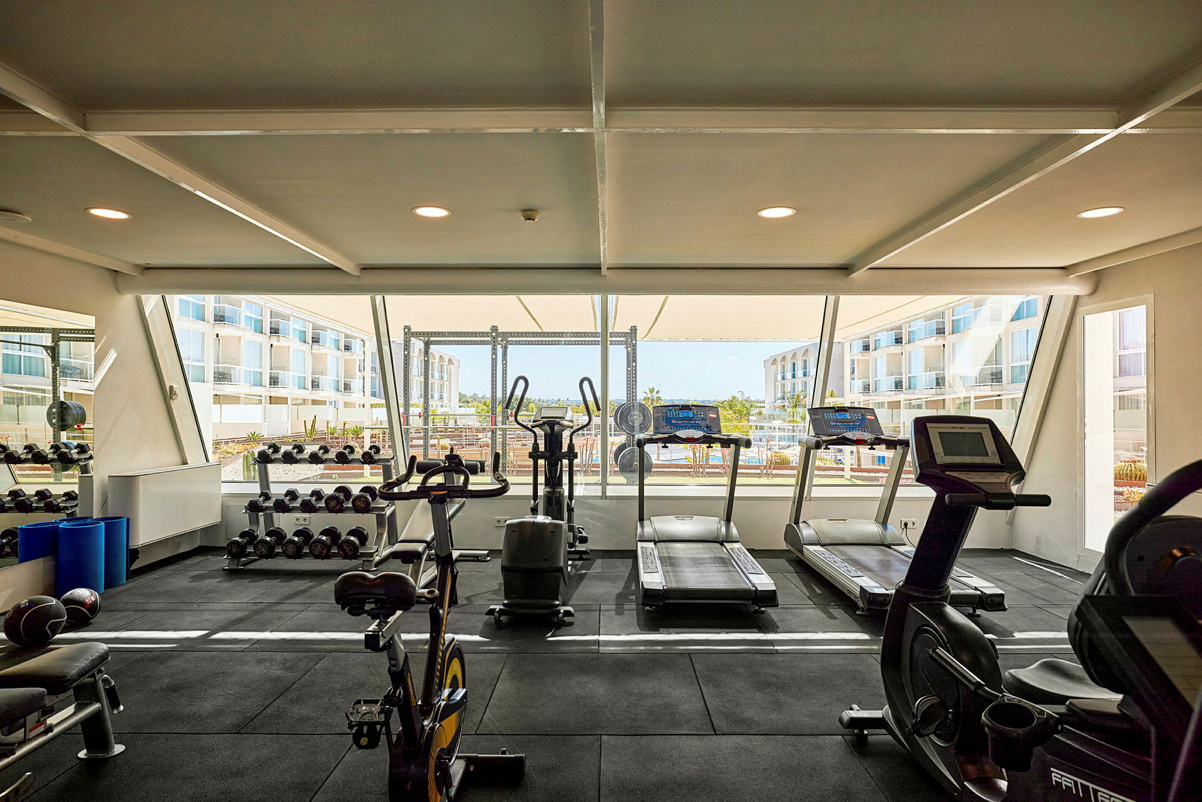 Fitnessrum med træningsmaskiner og vægte samt store vinduer med udsigt til omkringliggende bygninger. (AI-genereret tekst)