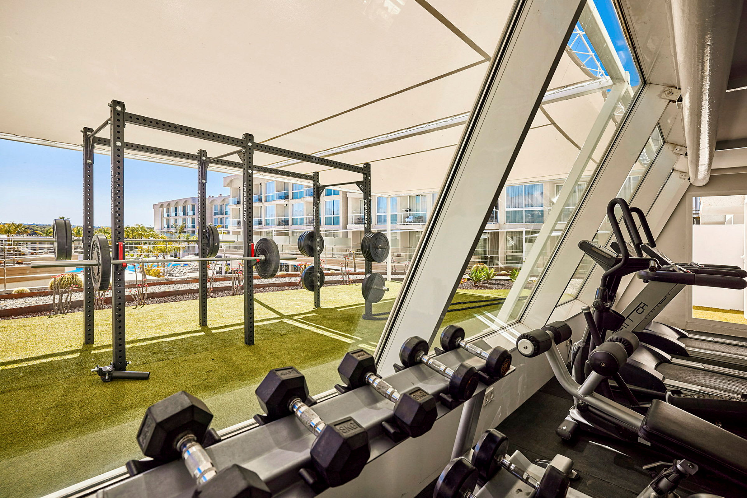 Et fitnesscenter med træningsudstyr som håndvægte og vægtløftningsstativer med udsigt over udendørsområdet. (AI-genereret tekst)
