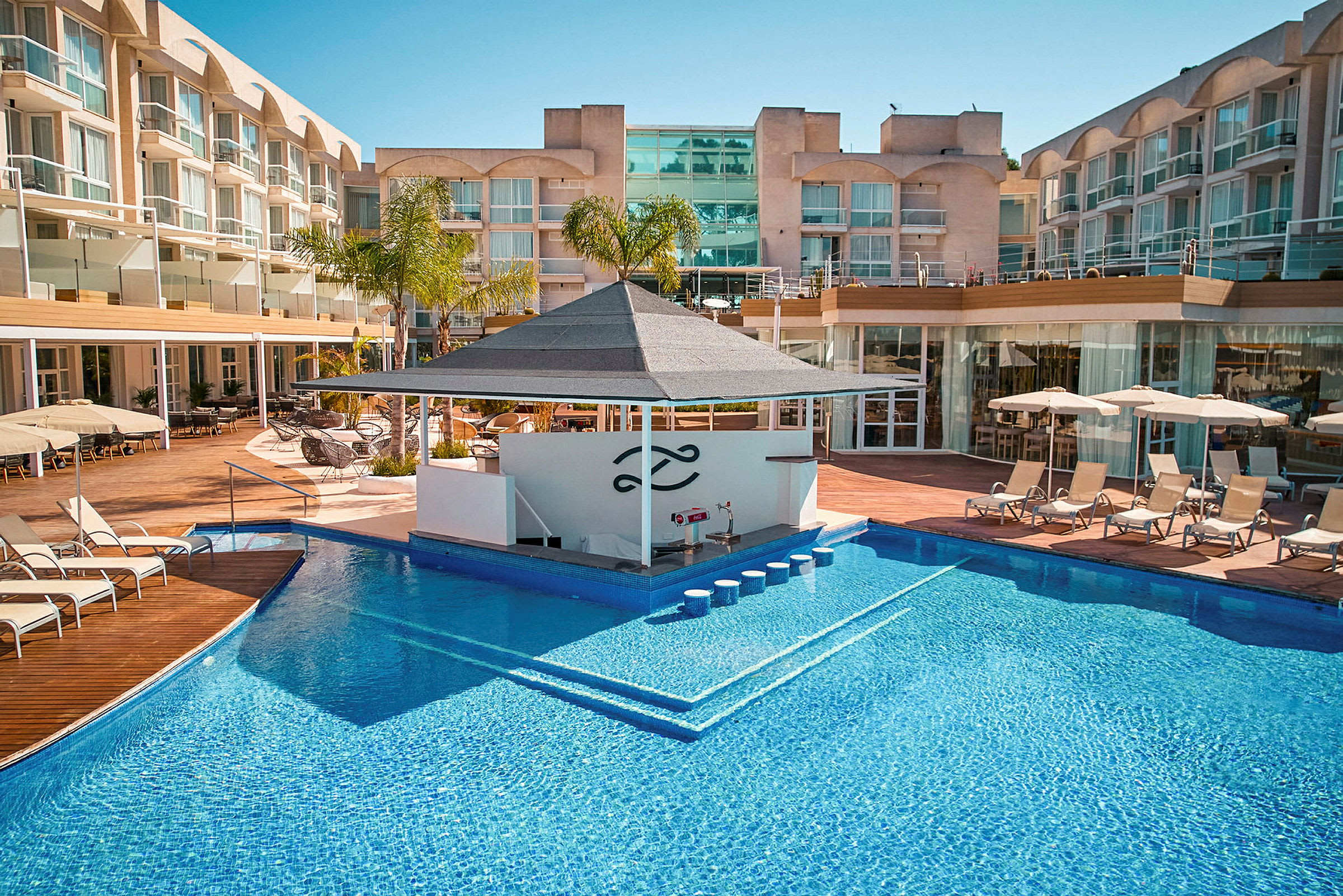Billede af et hotelområde med en udendørs pool, liggestole og en poolbar. (AI-genereret tekst)