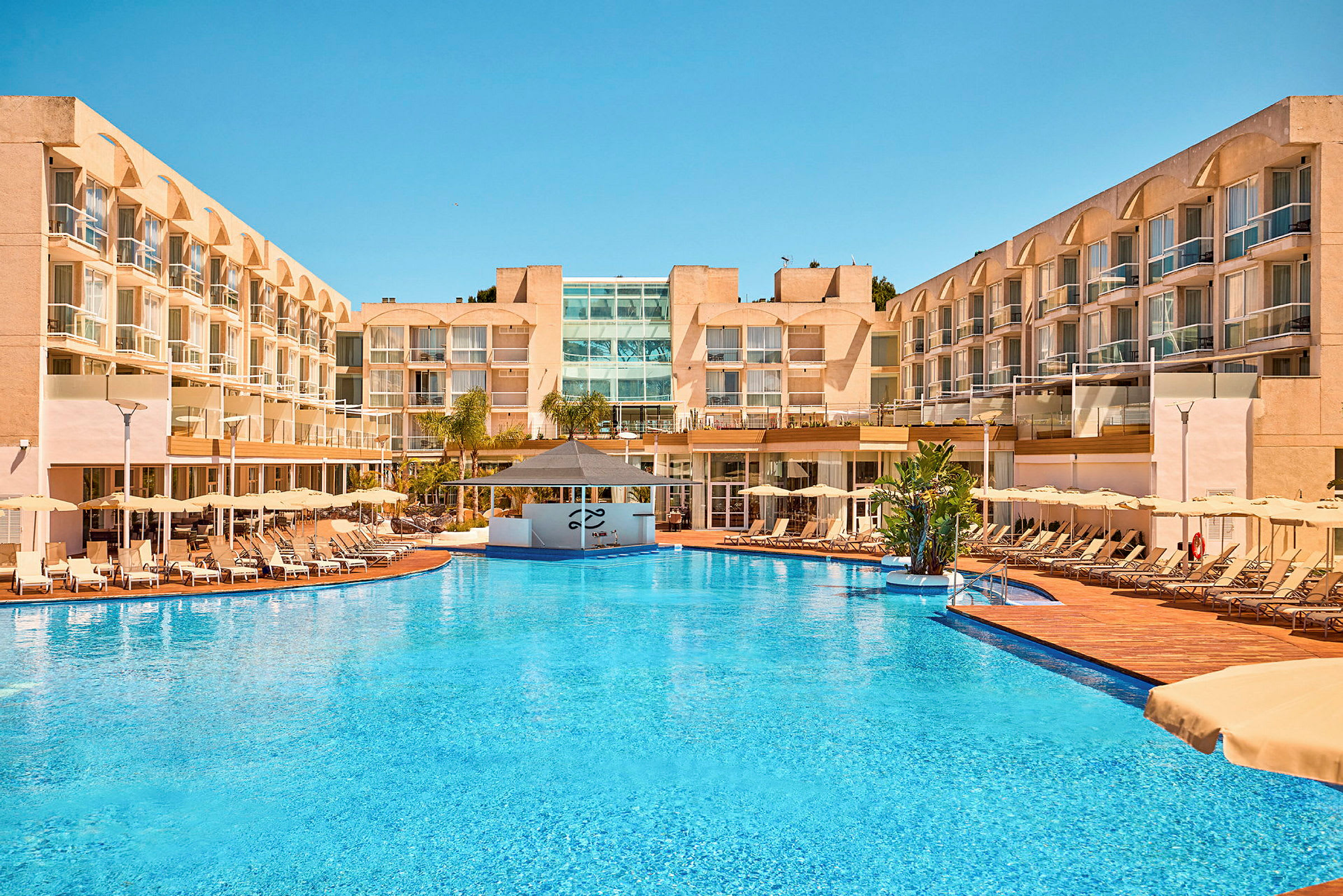 Et hotel med en stor udendørs pool, liggestole og parasoller omkring samt flere etager af altaner. (AI-genereret tekst)
