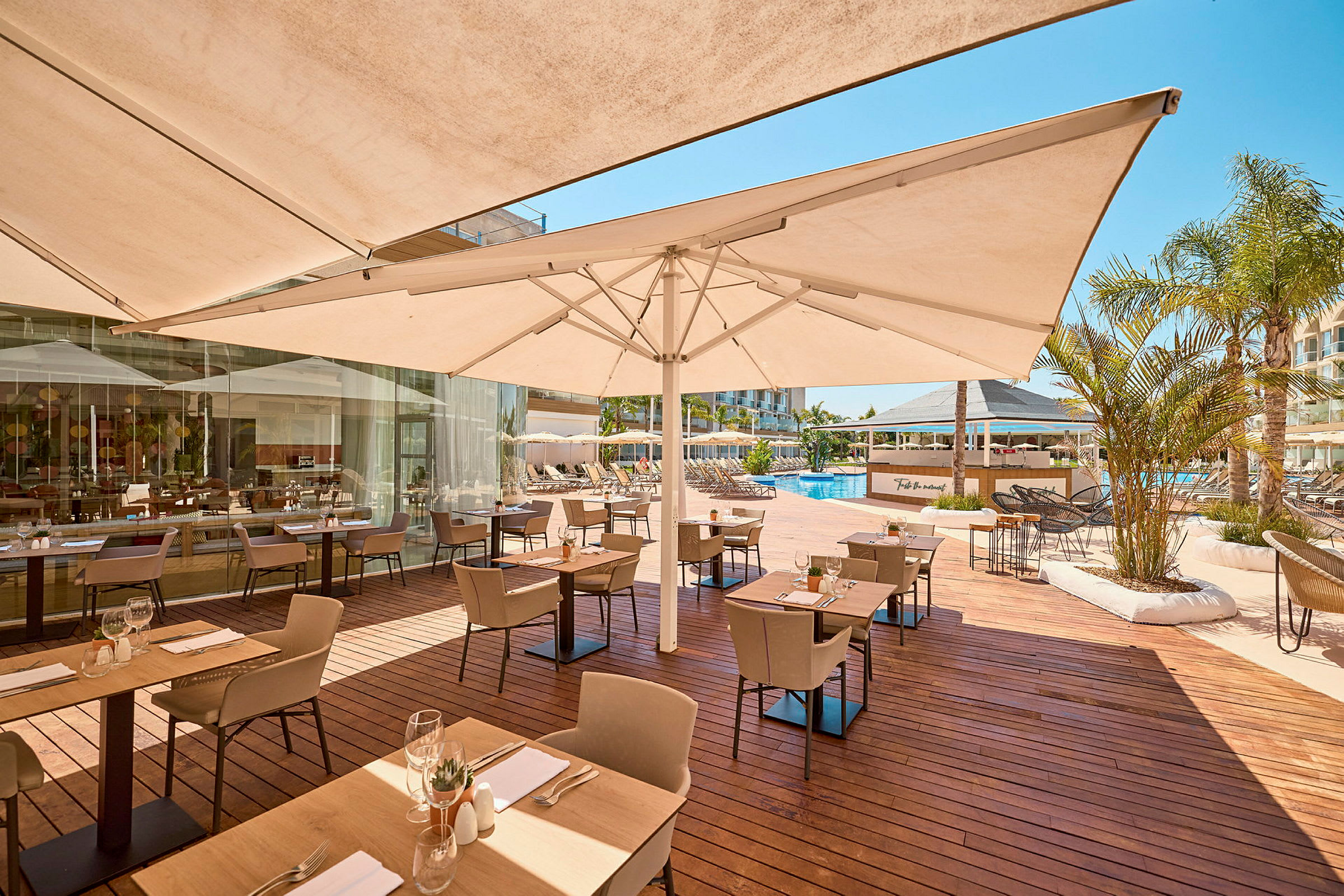 En udendørsrestaurant ved en pool, med borde og stole på en træterrasse under parasoller. (AI-genereret tekst)
