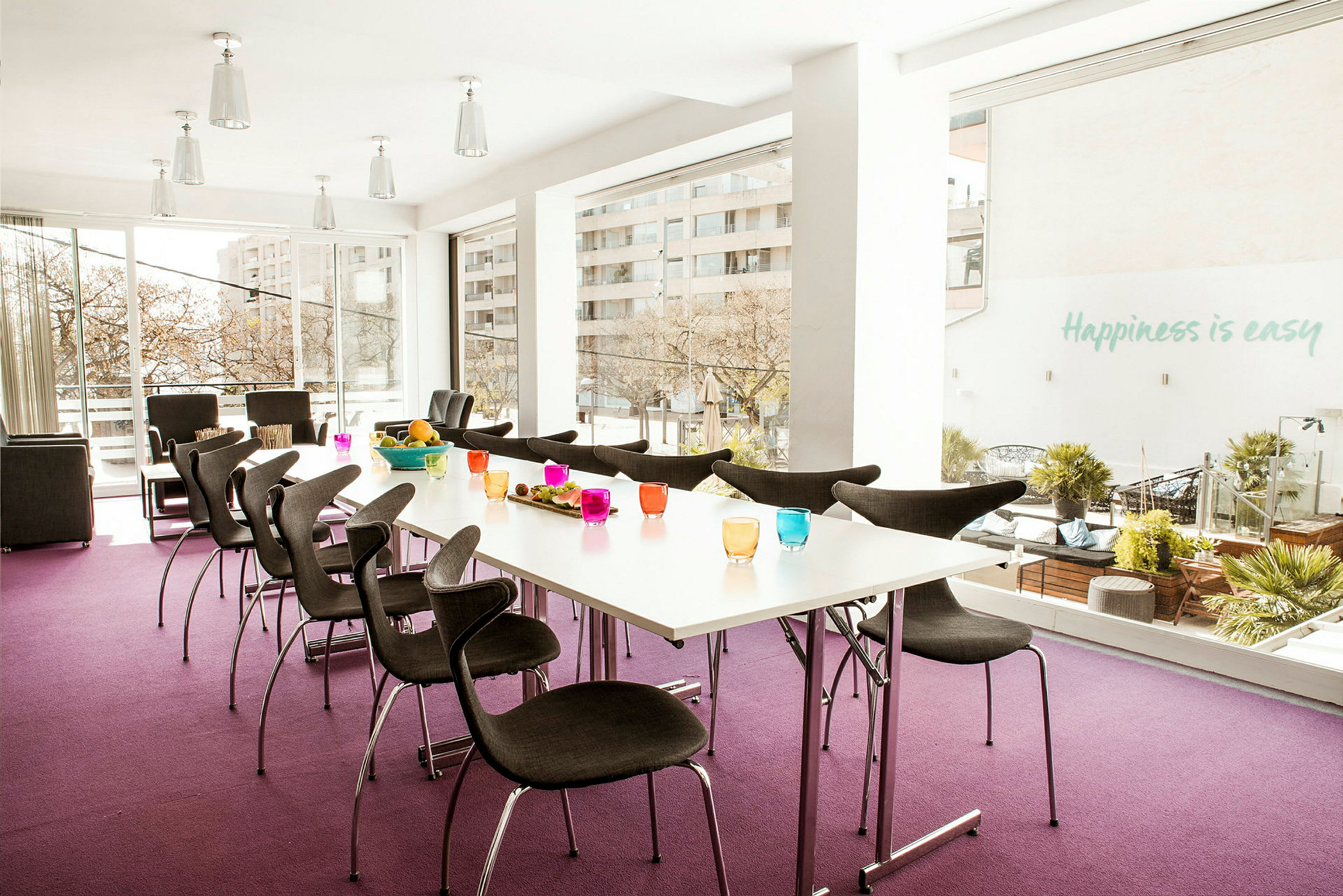 Et lyst, moderne konferencelokale med store vinduer, borde med farverige glas og mørke stole. (AI-genereret tekst)