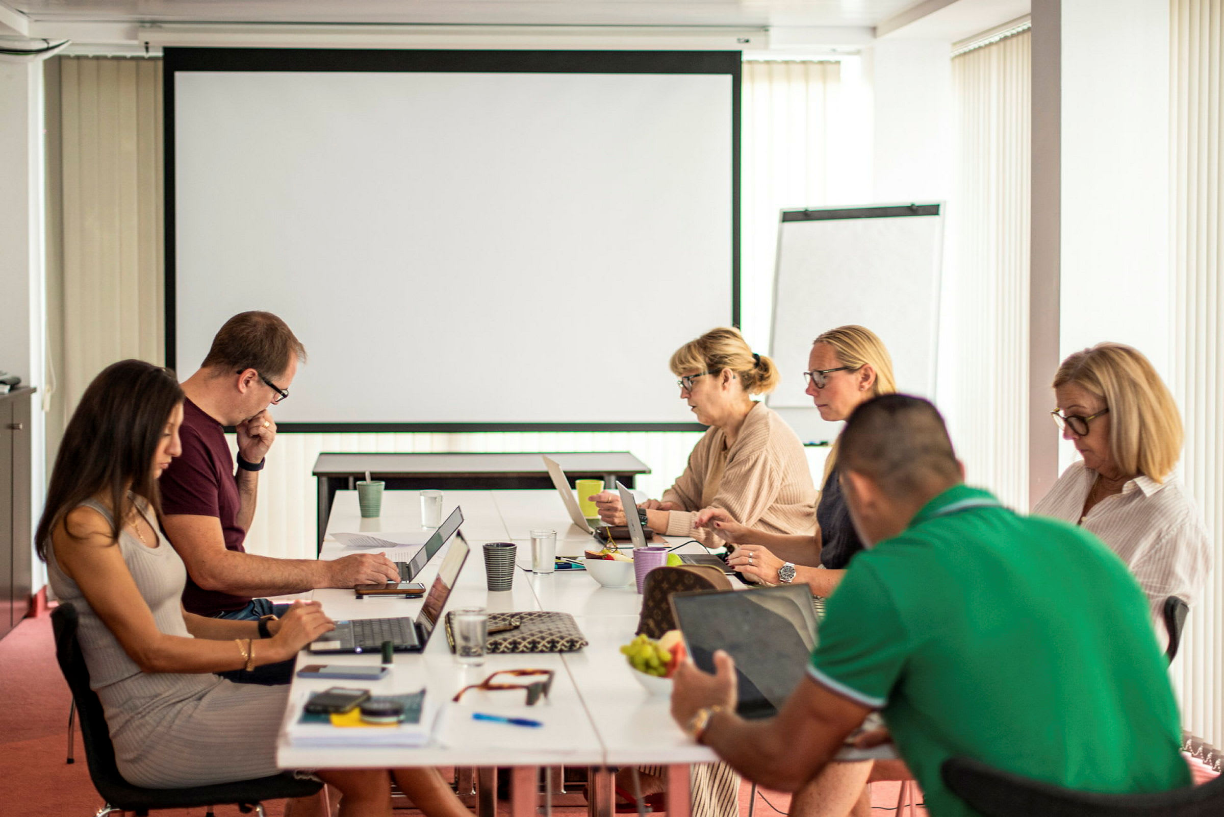 En gruppe mennesker sidder rundt om et bord med bærbare computere og notater i et mødelokale. (AI-genereret tekst)