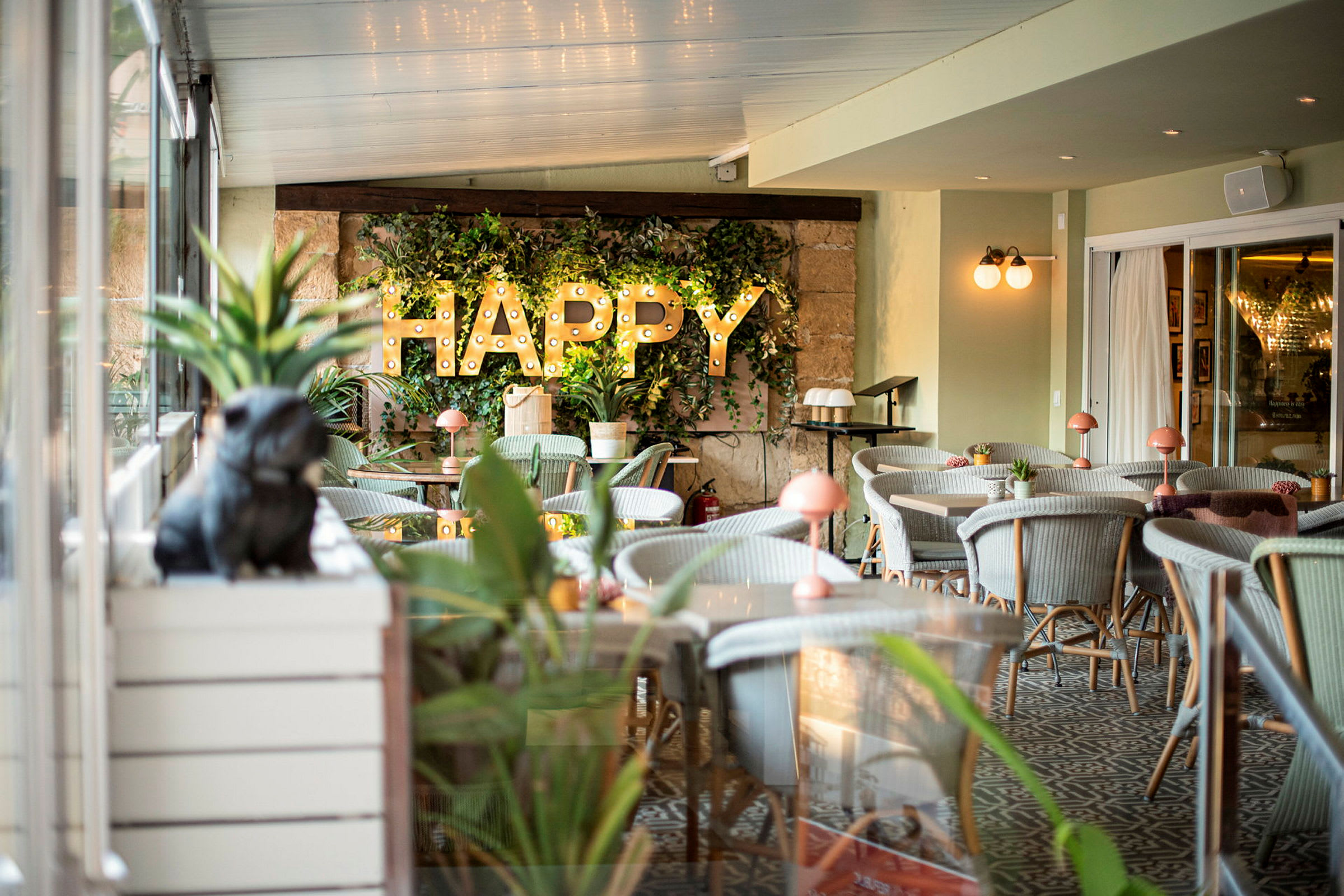 Interiør af en restaurant eller café med grønne planter, belysning og teksten 'HAPPY' på væggen. (AI-genereret tekst)