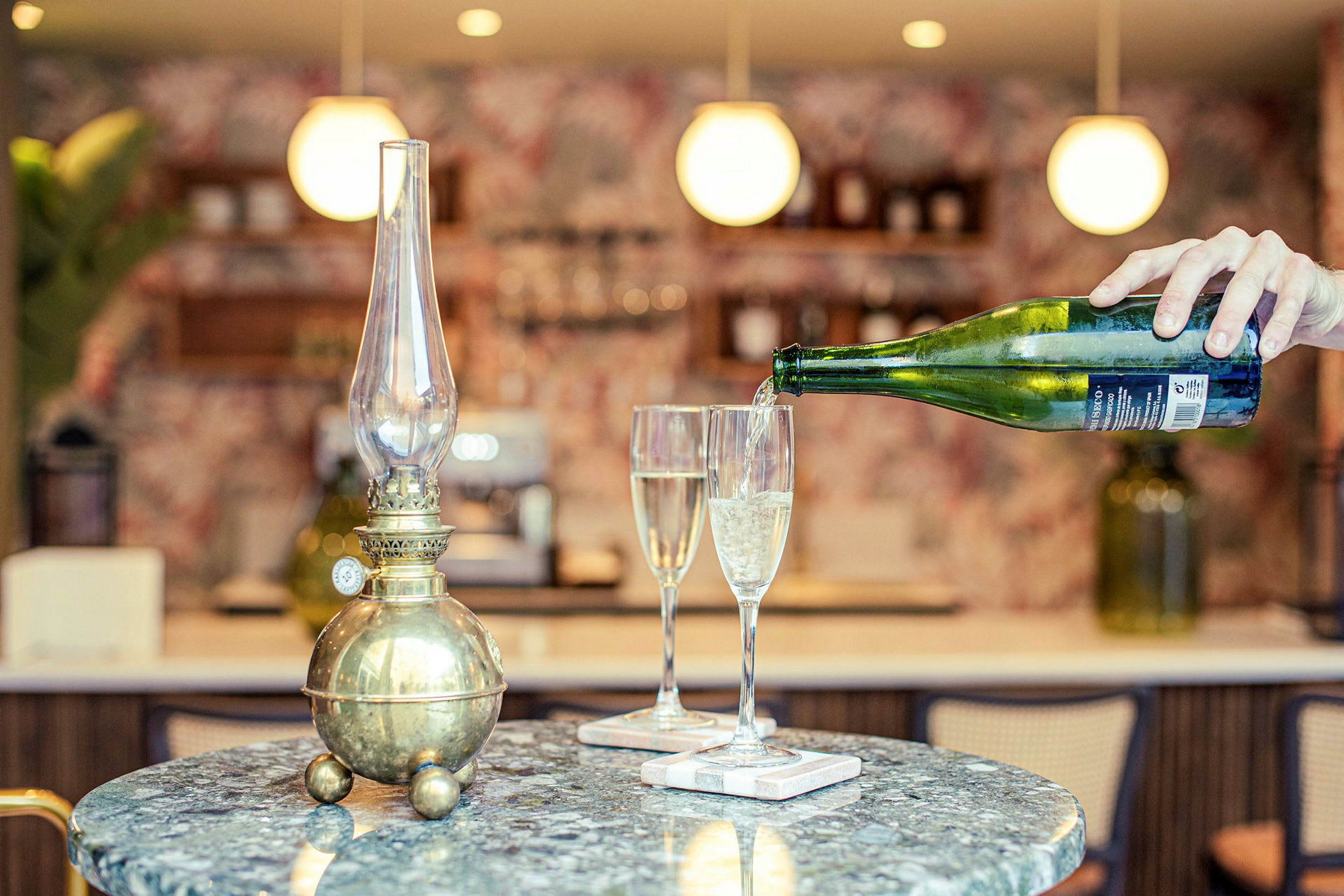 En antik olielampe på et bord ved siden af to champagneglas. En hånd hælder drikke i et af glassene. (AI-genereret tekst)