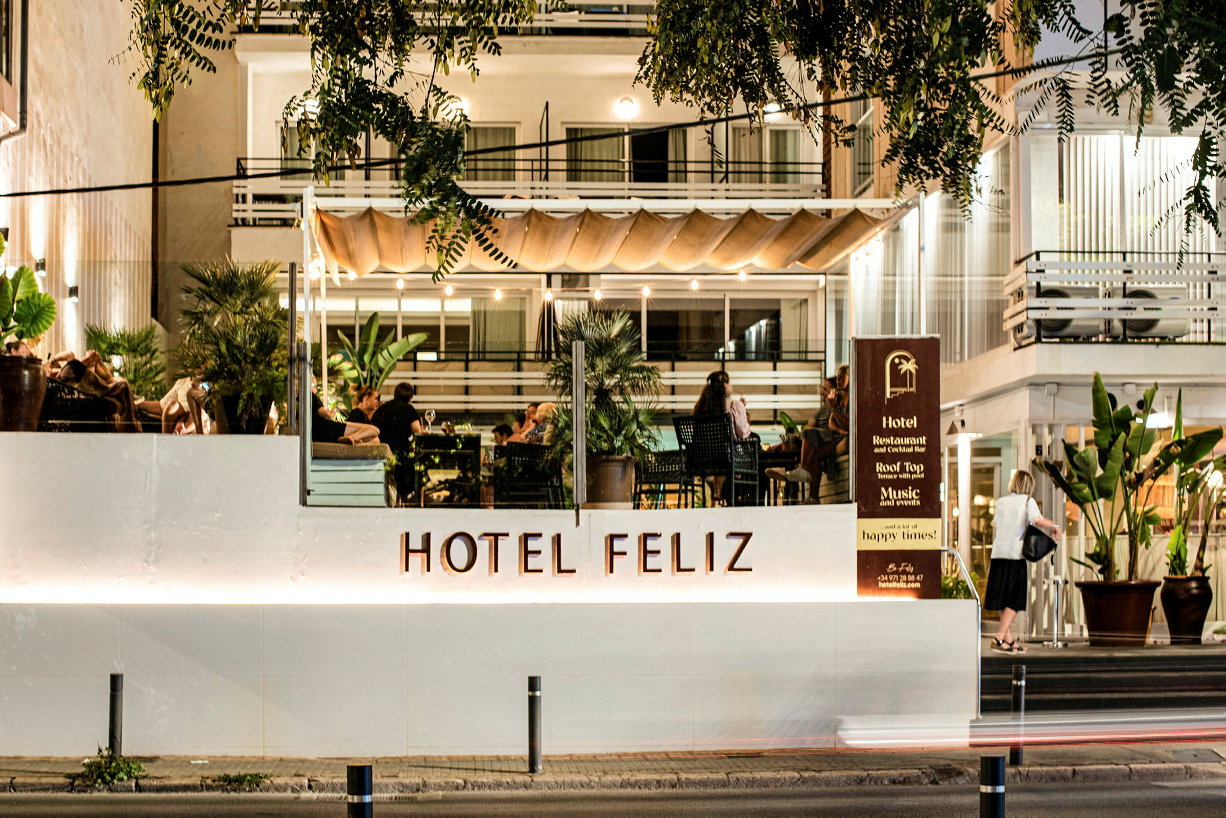 Hotel Feliz med oplyst facade og terrasse, hvor folk sidder. (AI-genereret tekst)