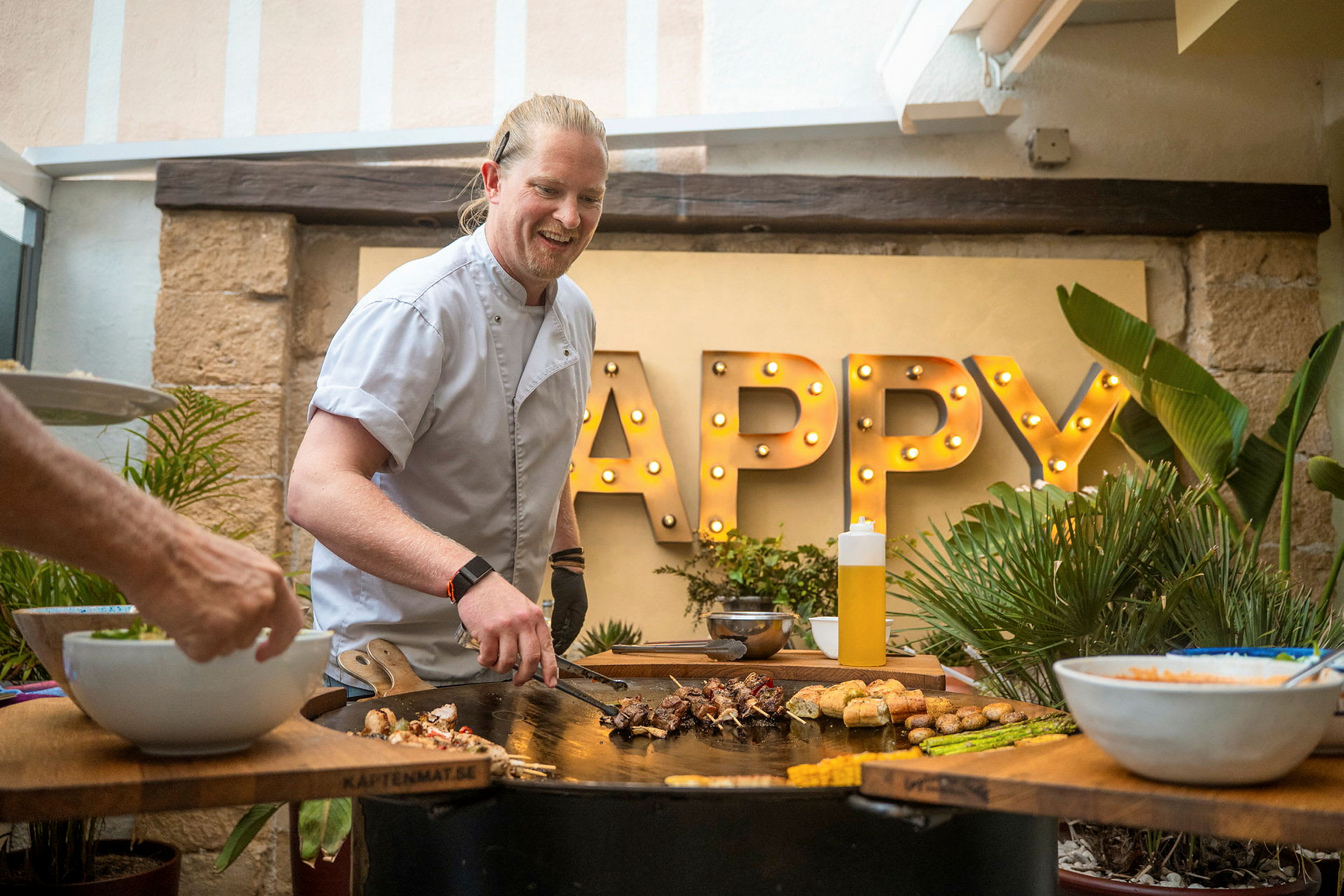 Person tilbereder mad på en grill med ordet 'Happy' oplyst i baggrunden. (AI-genereret tekst)