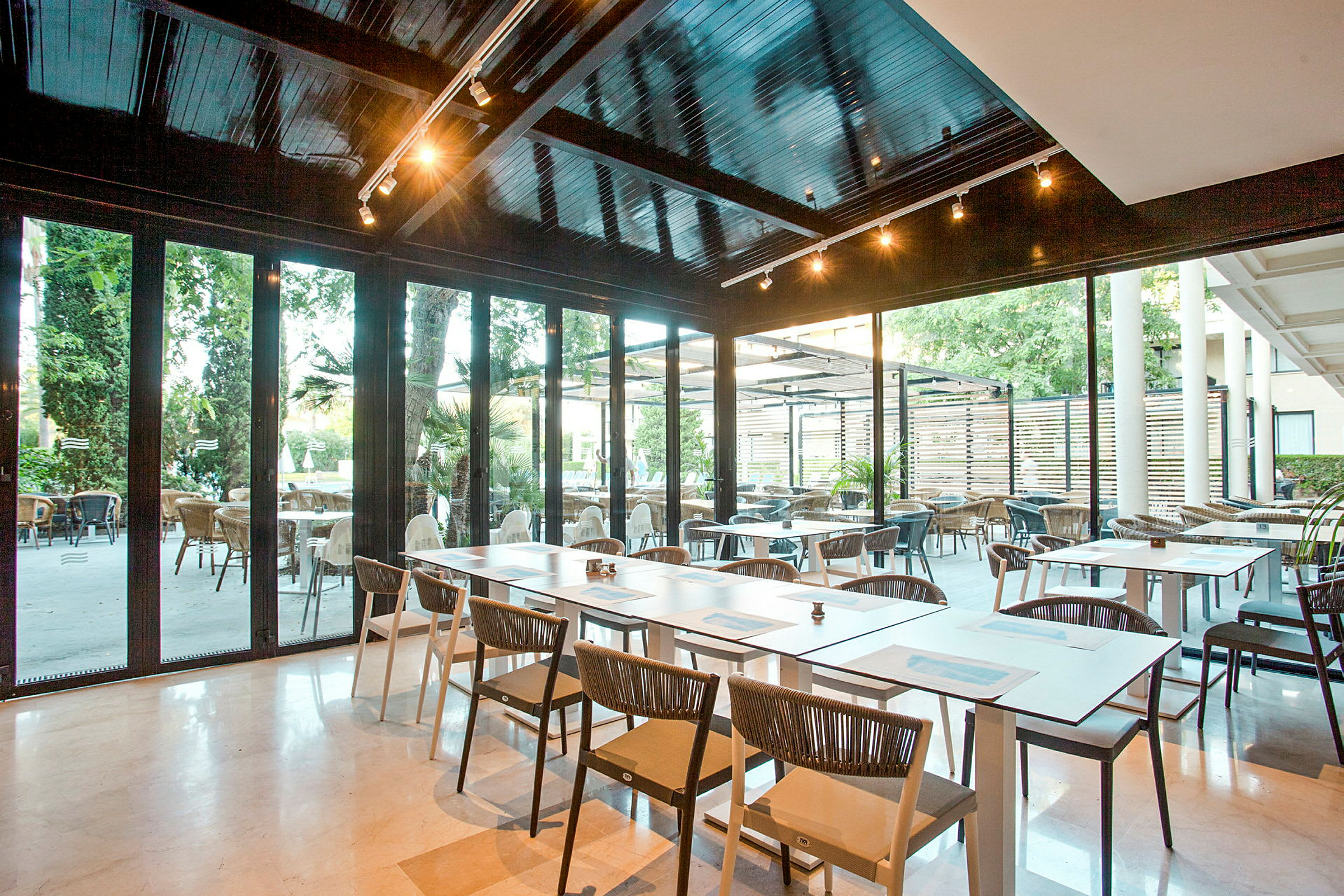 En moderne restaurant med siddepladser både indendørs og udendørs. Borde og stole er arrangeret på et lyst gulv, med glasvægge der giver udsigt til udendørsområdet. (AI-genereret tekst)