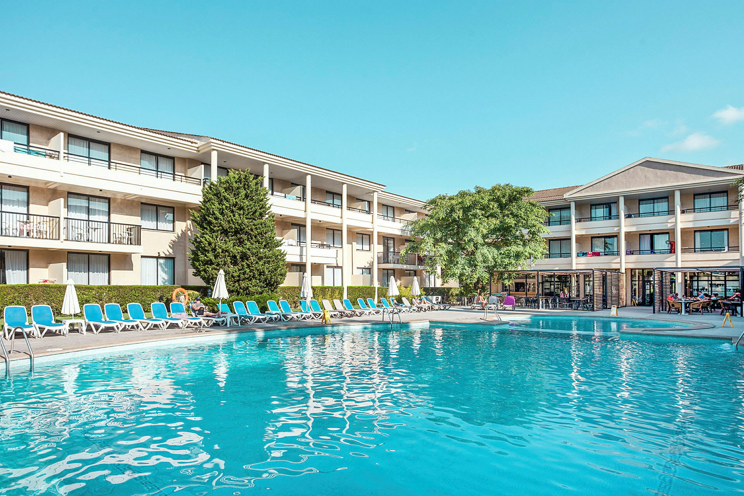Billede der viser et hotel med tre etager, liggestole og parasoller omkring en swimmingpool. Ved kanten er der en grøn plante og træer. (AI-genereret tekst)