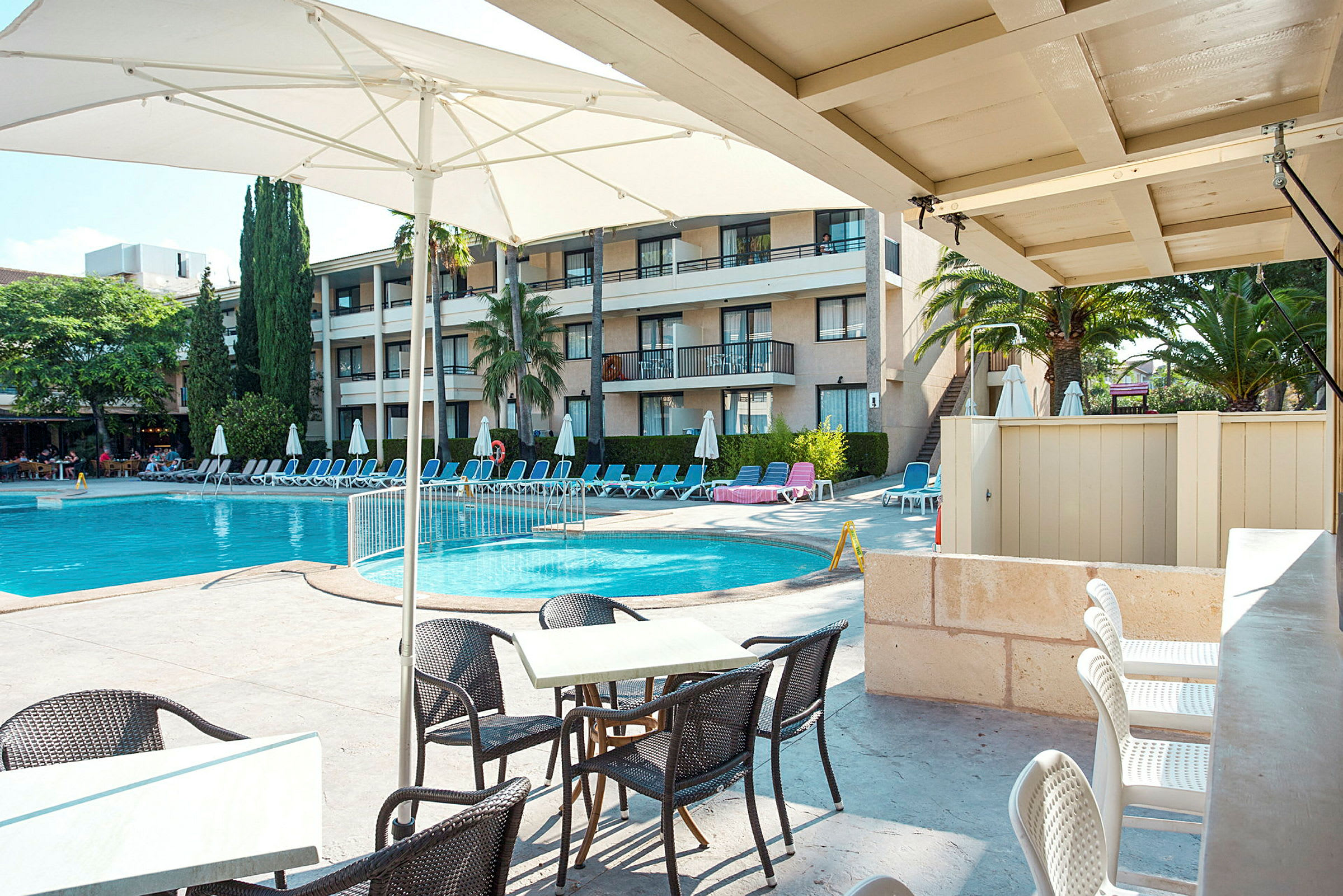 Poolområde med liggestole og parasoller ved en hotelbygning. I forgrunden er der borde og stole. (AI-genereret tekst)