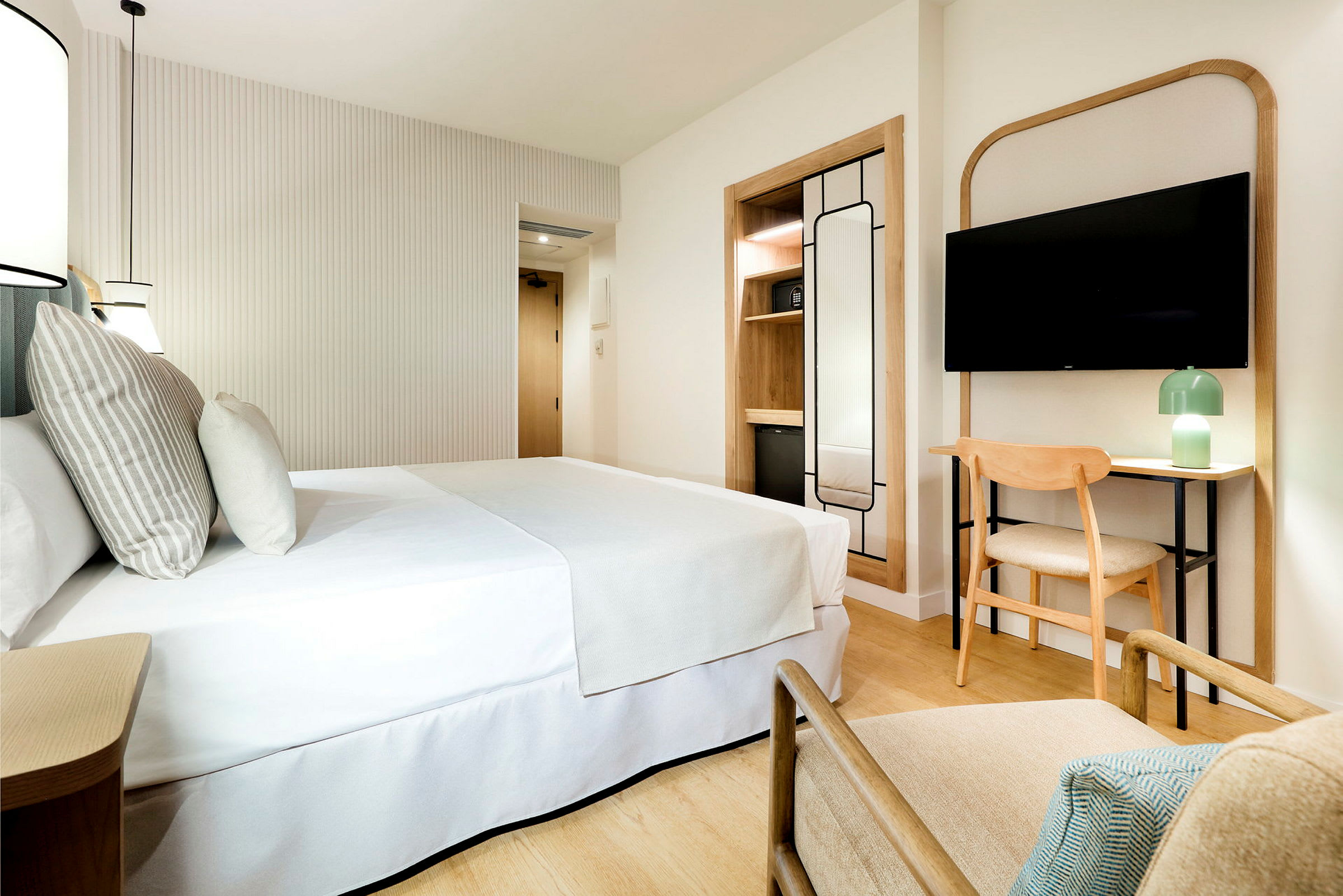Et moderne og lyst hotelværelse med en dobbeltseng, et TV på væggen, en lænestol, et skrivebord med stol og en minimalistisk indretning. (AI-genereret tekst)