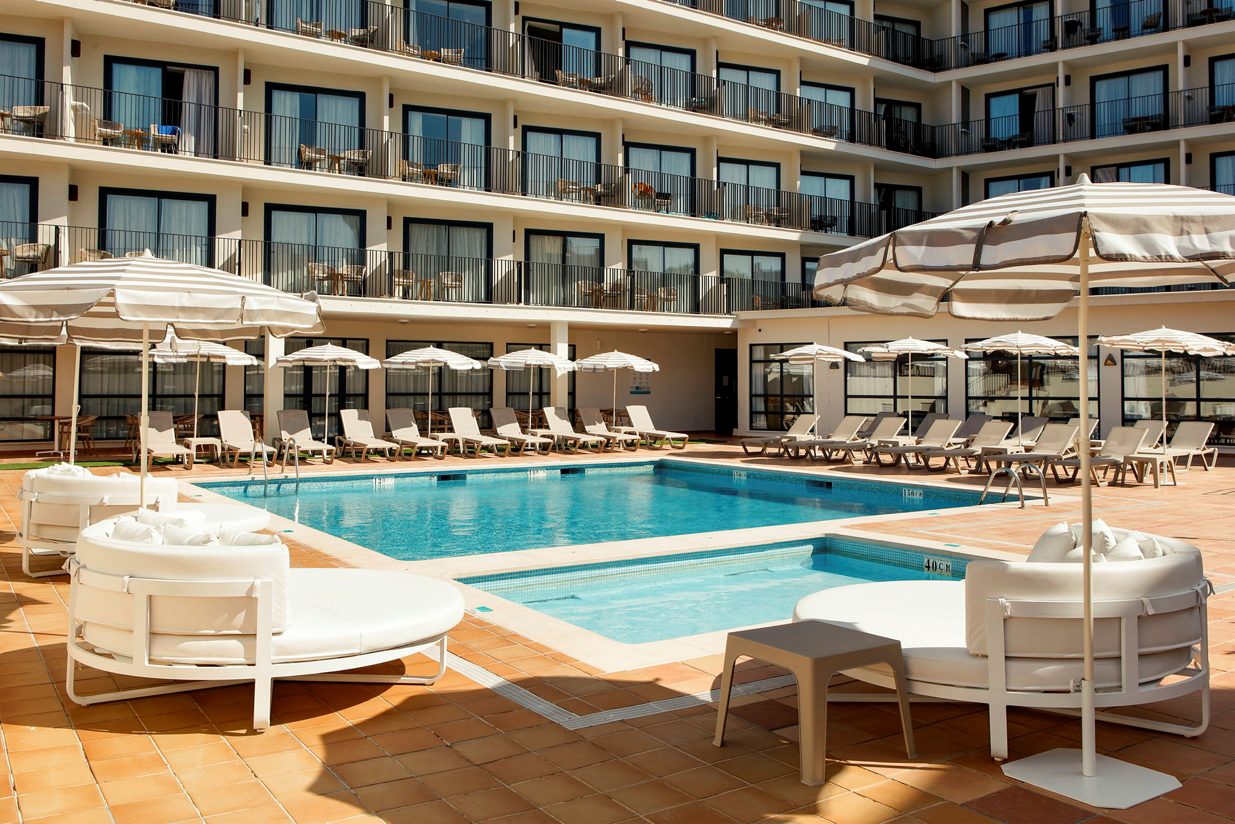 Et hotelpoolområde med liggestole, parasoller og en pool omgivet af flere etager med altaner. (AI-genereret tekst)
