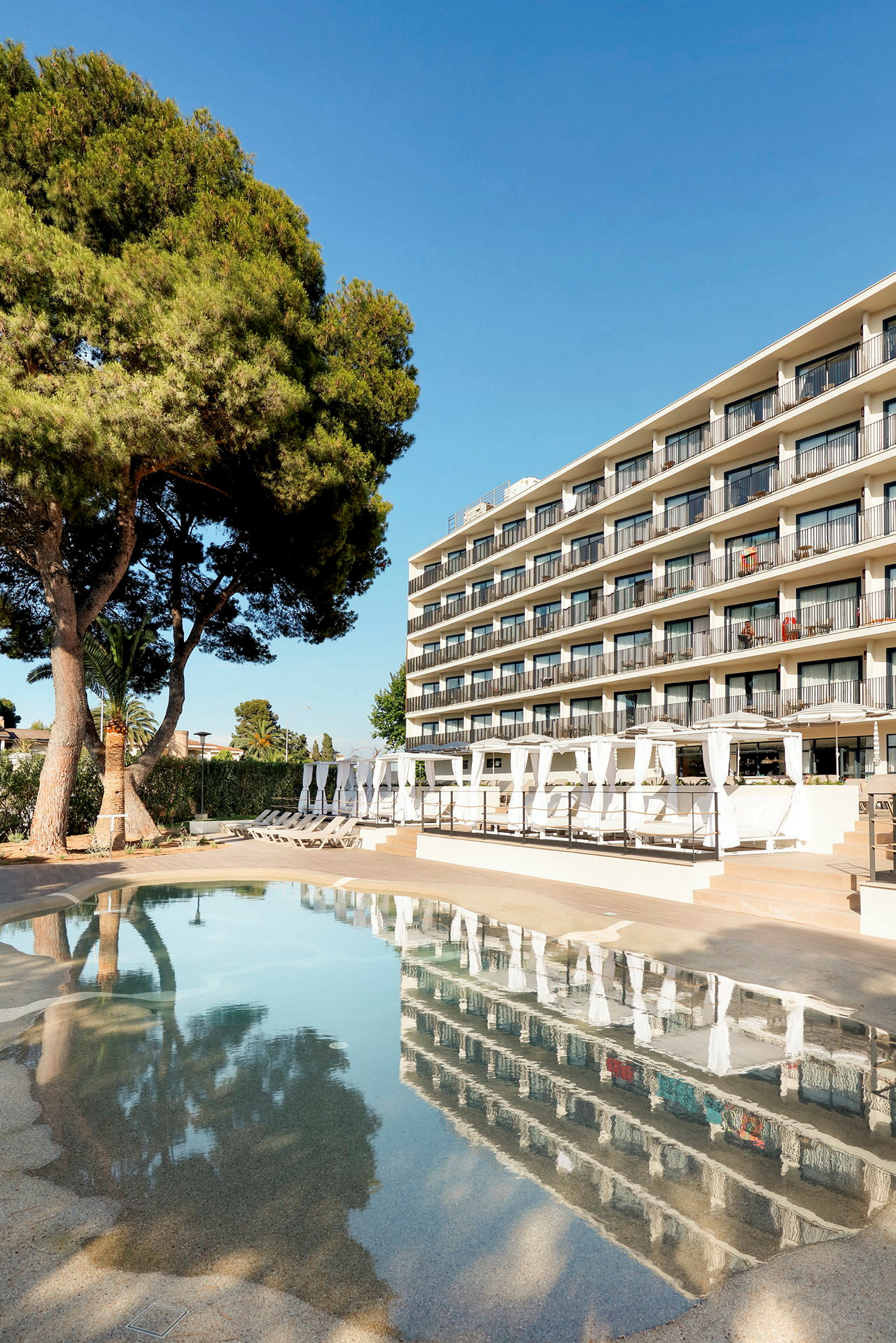 Et hotel med flere altaner ved en pool og træer i forgrunden. (AI-genereret tekst)