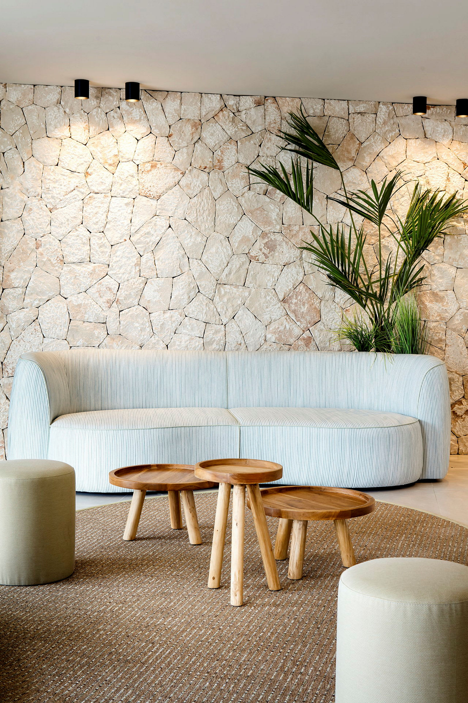 En lys stueindretning med en beige sofa, små træborde og dekorative planter foran en væg med stenstruktur. (AI-genereret tekst)