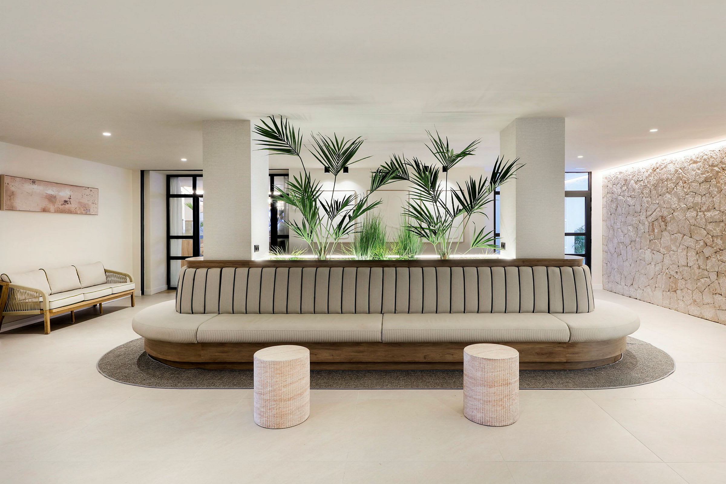 En moderne ventesal eller lounge med en lang, polstret sofa, to runde skamler foran og grønne planter som dekoration i baggrunden. (AI-genereret tekst)