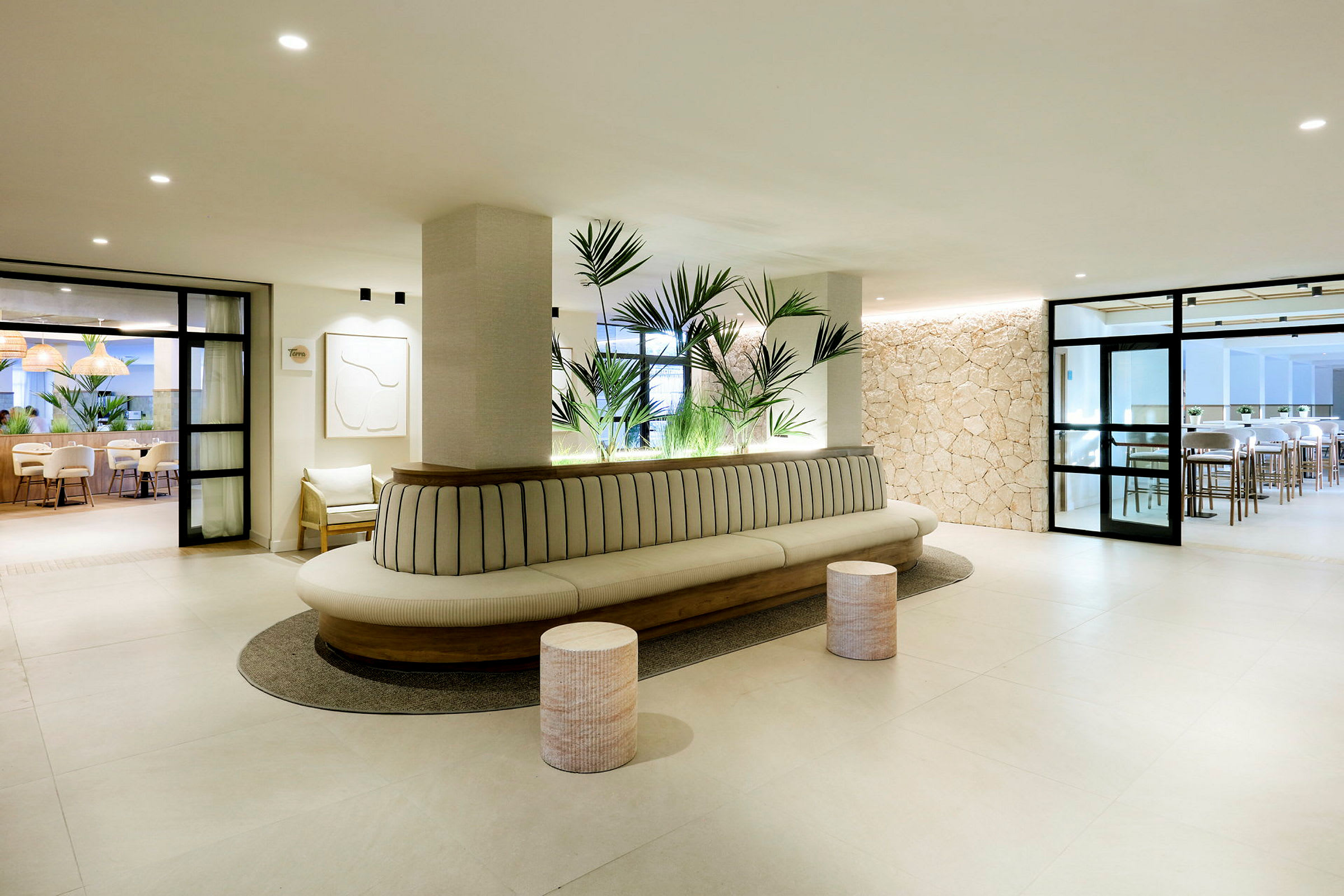 En moderne lobby med lys indretning, en lang sofa med planter bagved og stole samt borde ses i baggrunden. (AI-genereret tekst)