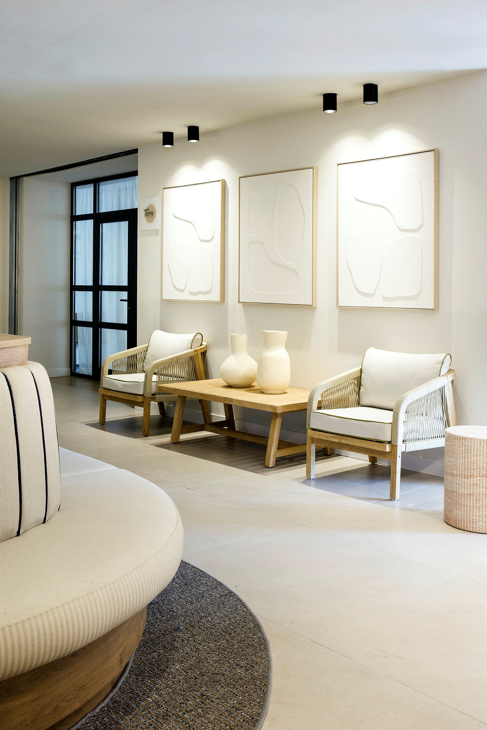 Et moderne og elegant loungeområde med to lænestole, et træbord med to dekorative vaser og tre minimalistiske malerier på væggen. (AI-genereret tekst)