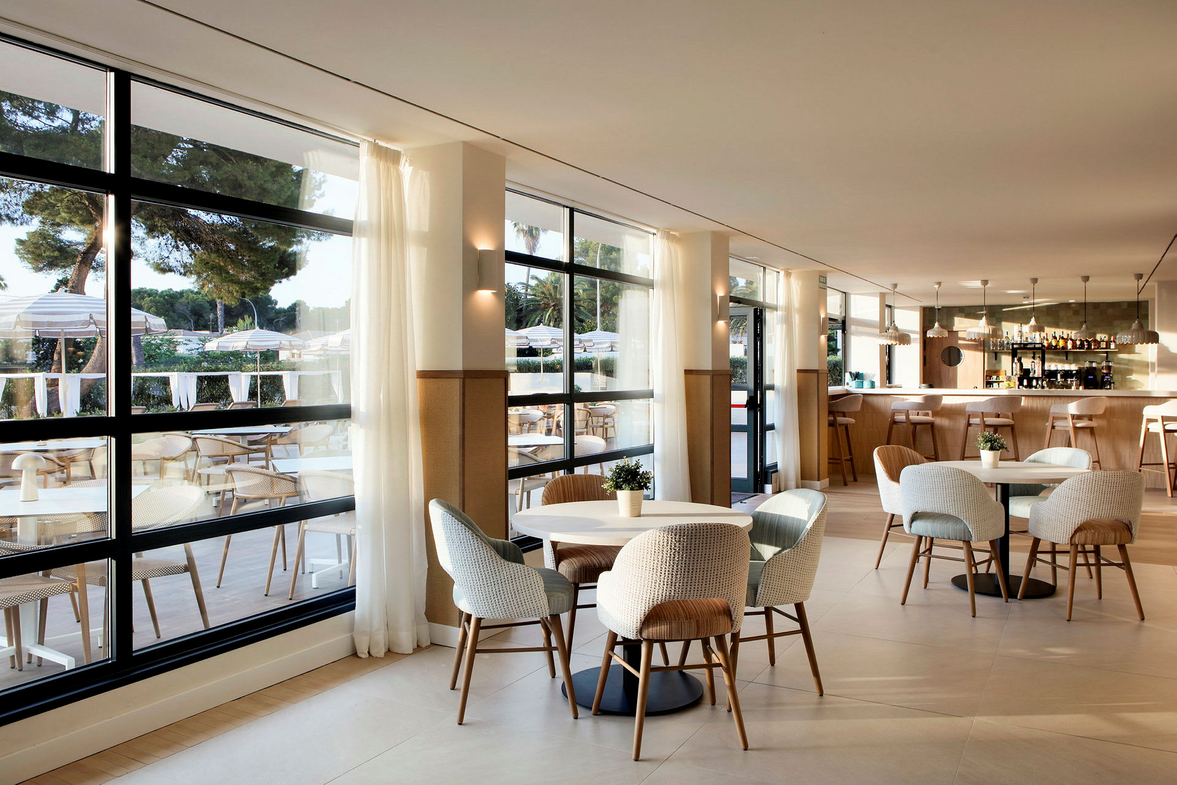 En moderne restaurant med store vinduer, lyse møbler og en indbydende indretning. Udsigt til en terrasse med borde og parasoller. (AI-genereret tekst)