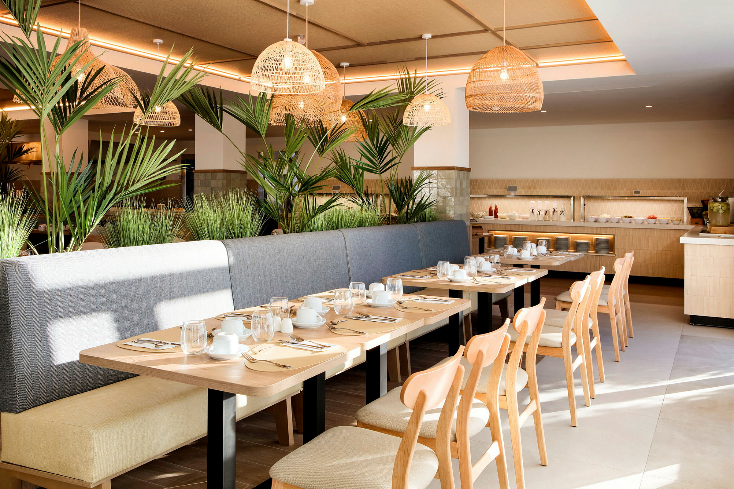 En lys og moderne restaurant med træborde, stole og bænk af stof. Dekoreret med grønne planter og flettede loftslamper. Porcelæn og glas er dækket på bordene. (AI-genereret tekst)