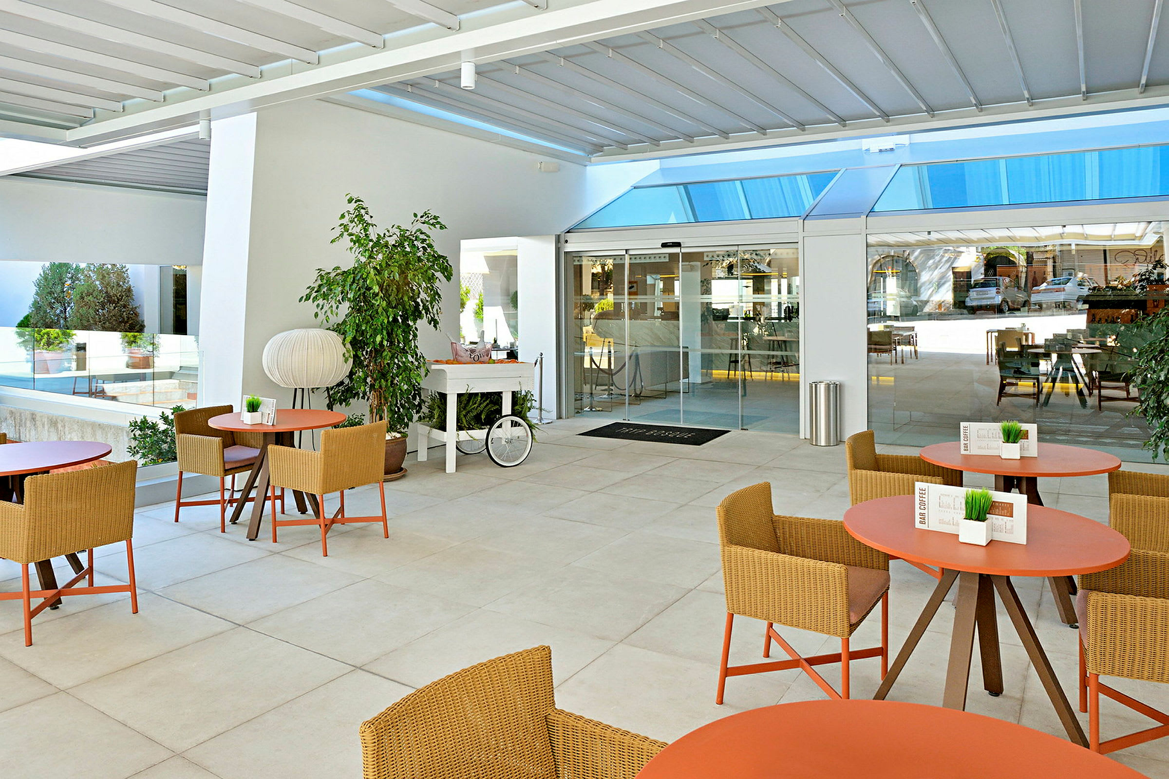 Billedet viser en moderne udendørs terrasse med borde og stole i rattan-design, omgivet af grønne planter og med glaspartier i baggrunden. (AI-genereret tekst)