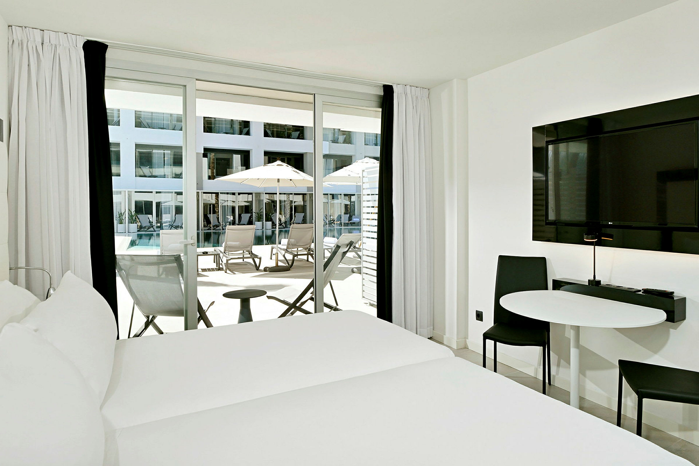 Hotelværelse med seng, bord, stol og altandør med adgang til en terrasse med liggestole og parasoller ved en pool. (AI-genereret tekst)