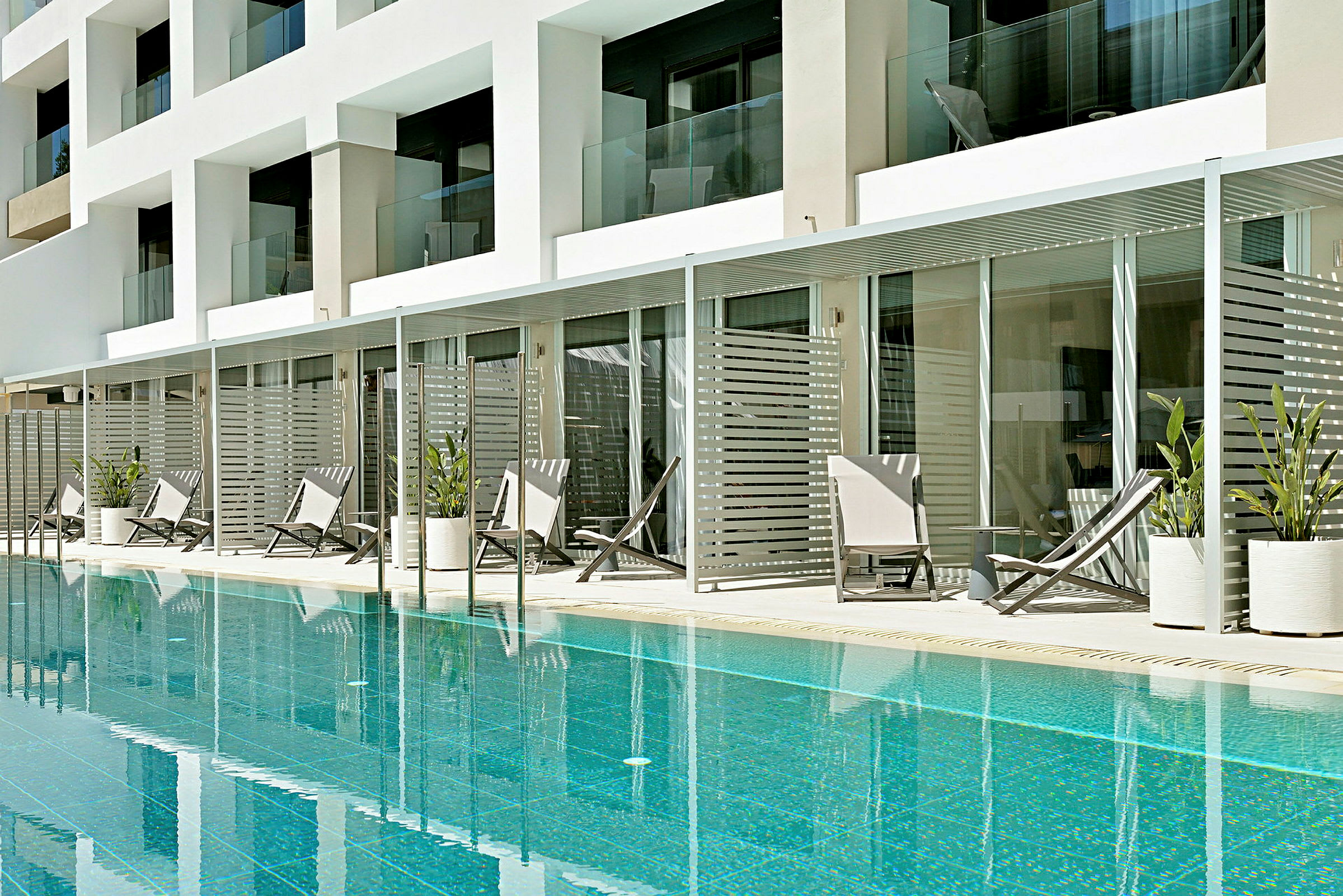 En swimmingpool foran en moderne hotelbygning med store altaner og liggestole omgivet af plantede planter. (AI-genereret tekst)