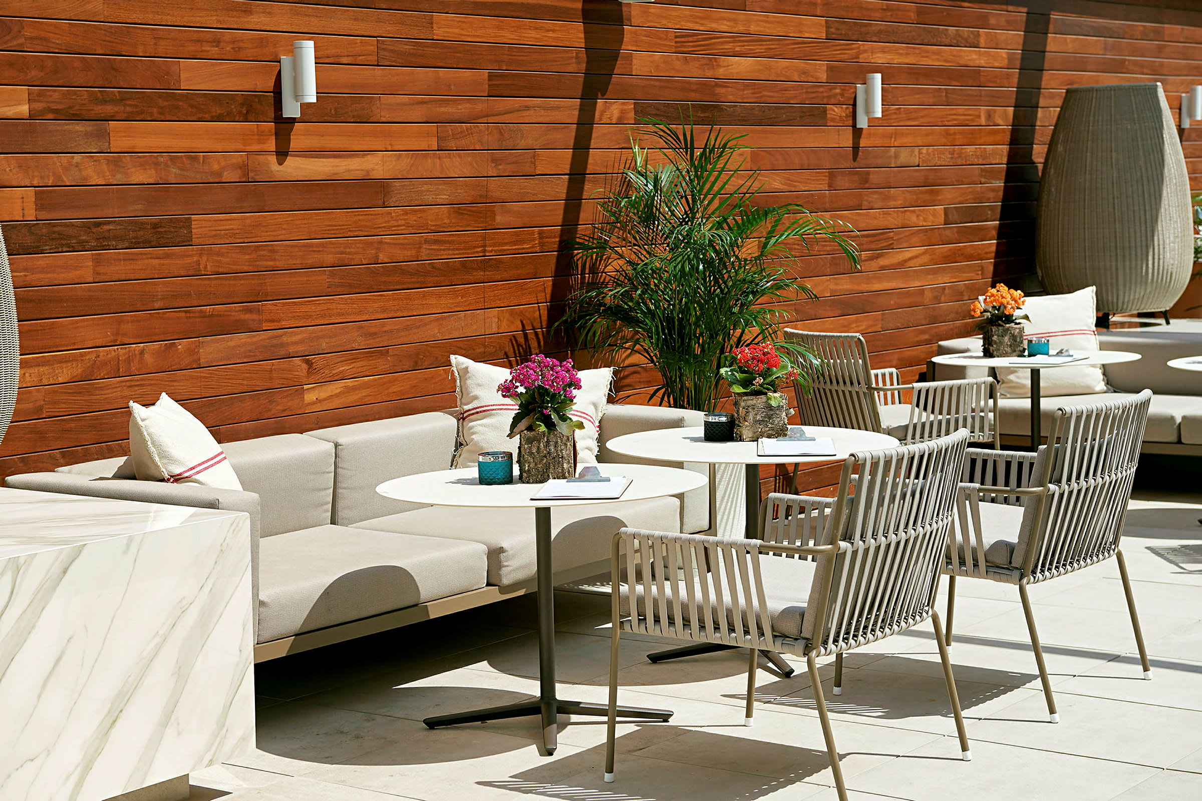 Et stilfuldt møblement på en terrasse med borde og stole, pyntet med planter og puder, mod en træpanelvæg. (AI-genereret tekst)