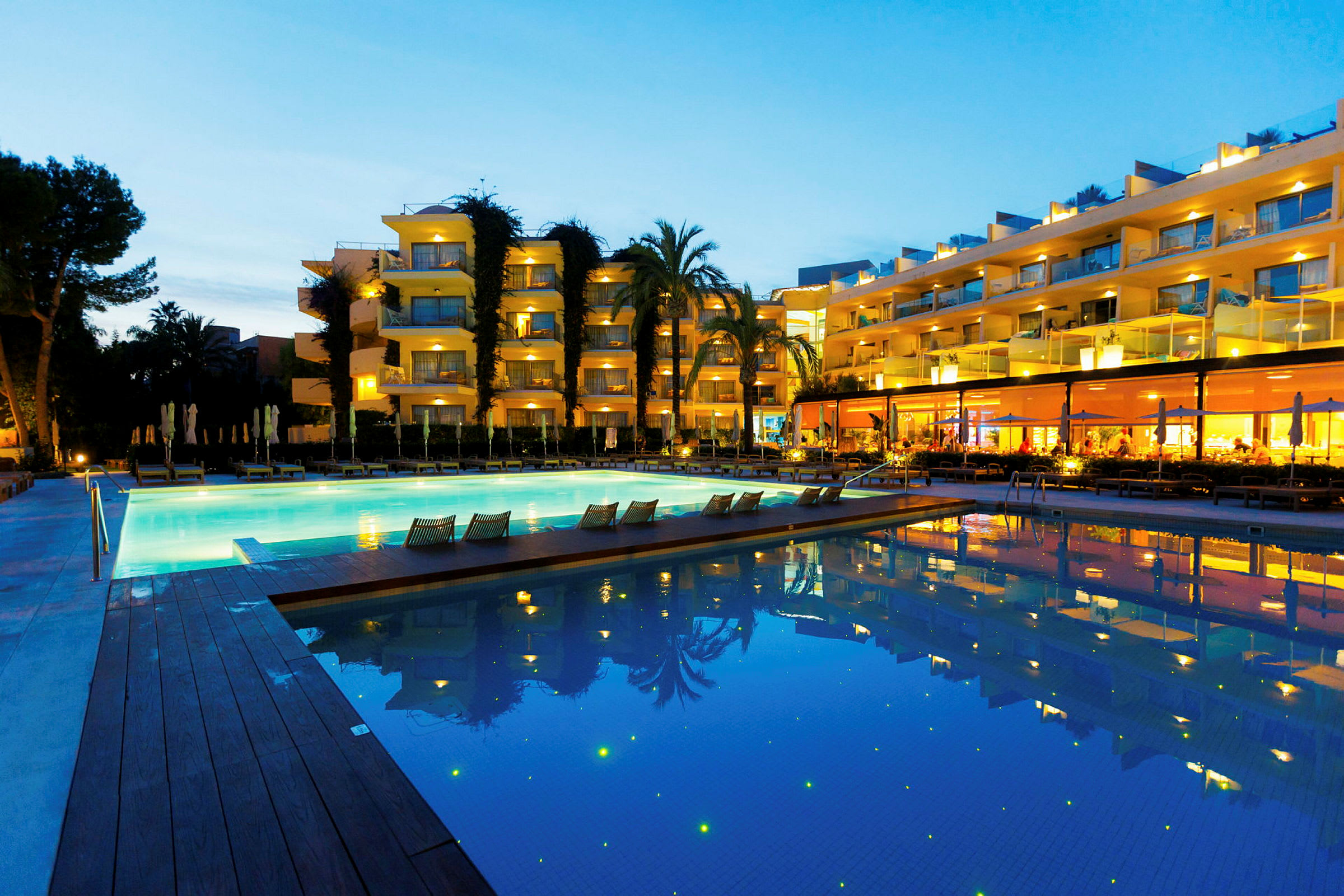 Hotel med lyse facader, en swimmingpool og omkringliggende grønne områder om aftenen. (AI-genereret tekst)
