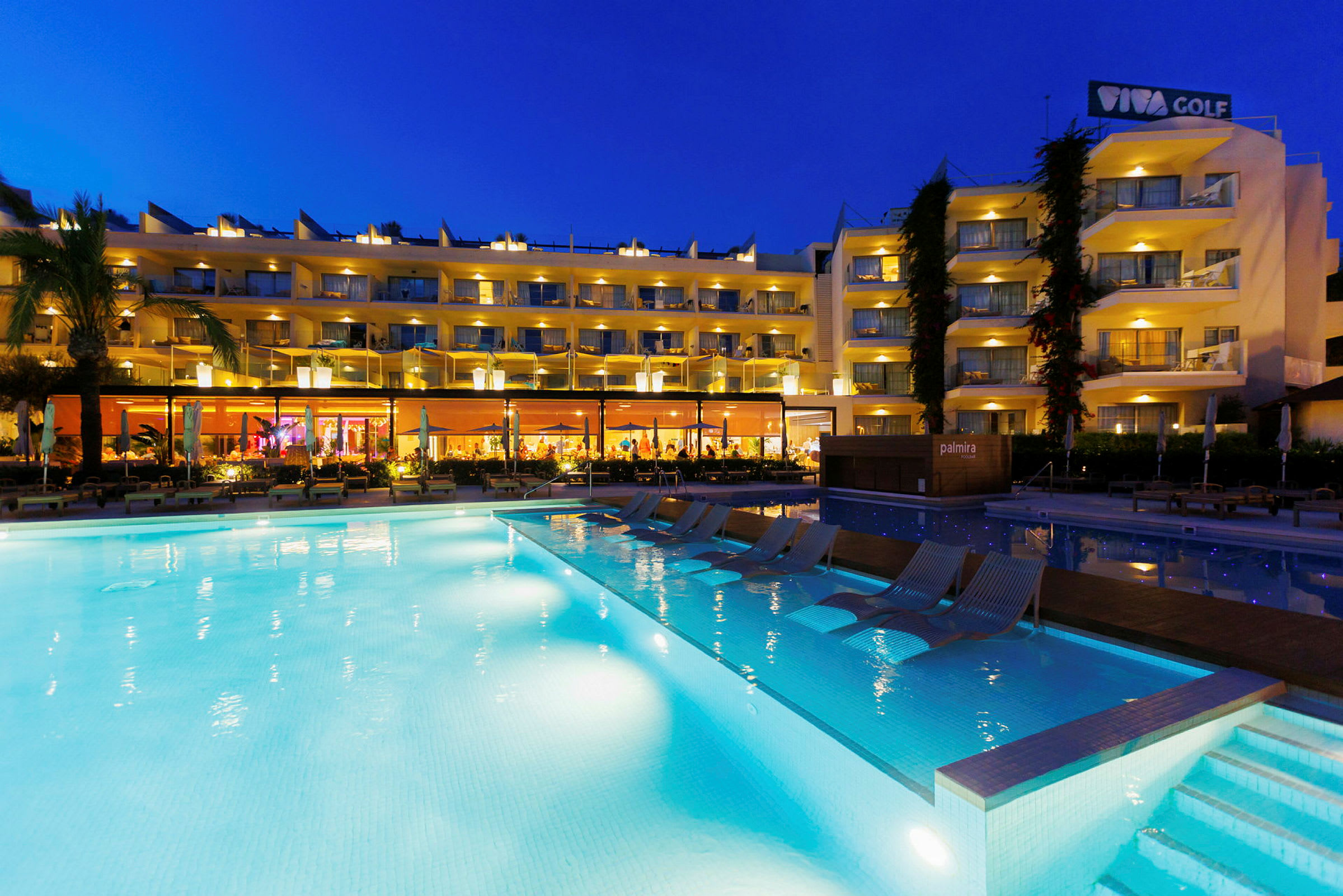 Hotelbygning med oplyste altaner og en belyst pool i forgrunden om aftenen. (AI-genereret tekst)