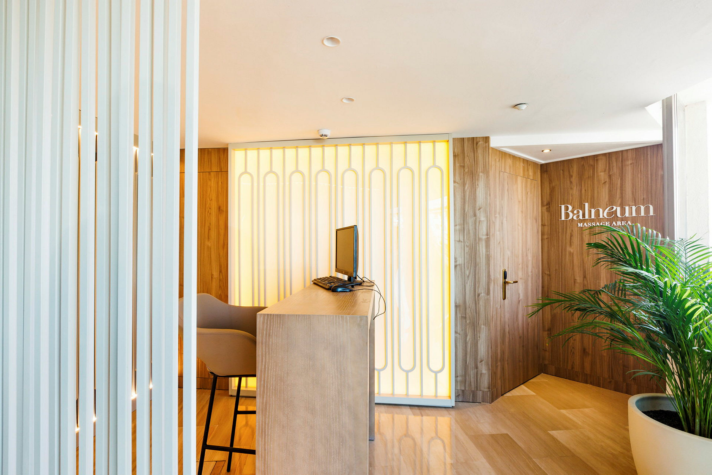 Reception med bord, computer og stol, med en baggrund af vægpaneler og en dør mærket 'Balneum Massage Area'. (AI-genereret tekst)