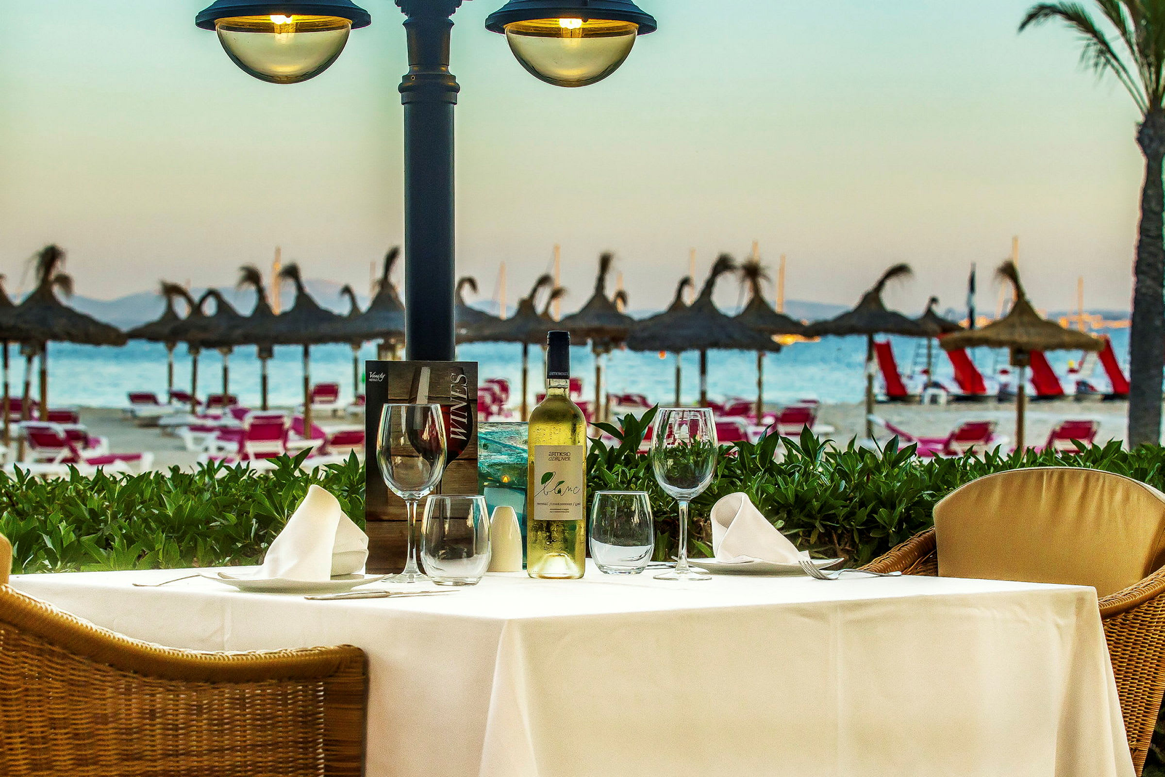 Et bord med opdækning og en vinflaske står på en terrasse med udsigt over stranden og parasoller. (AI-genereret tekst)