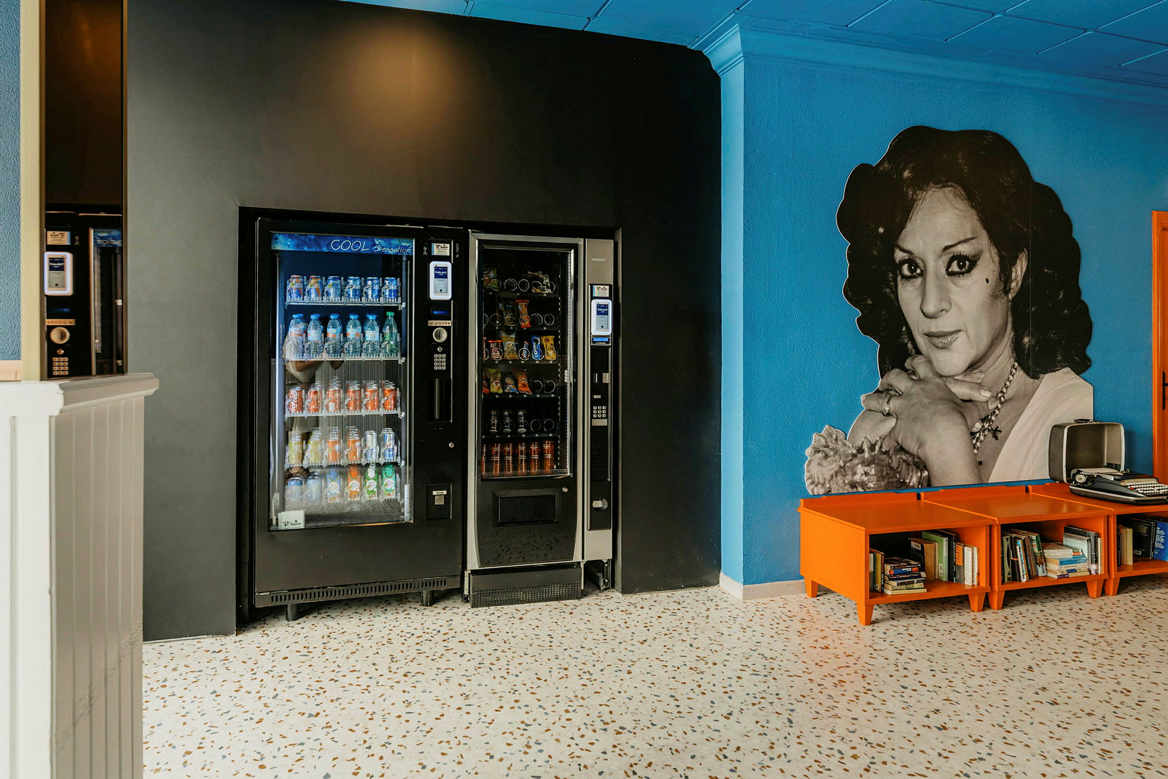 Billede af to automater til drikkevarer og snacks placeret mod en sort væg, sammen med et område med orange hylder indeholdende bøger foran en blå væg. (AI-genereret tekst)