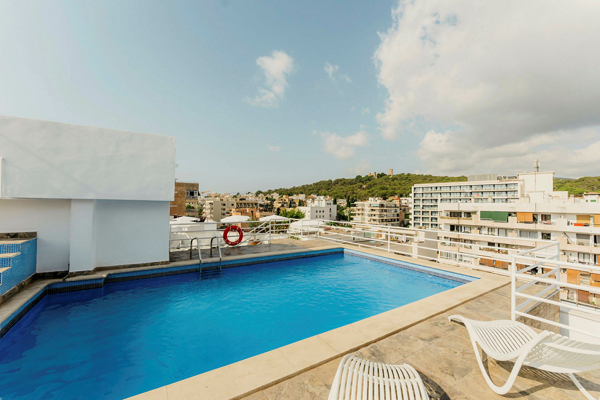 En tagterrasse med en pool, liggestole og udsigt over byen omgivet af bygninger og grønne områder. (AI-genereret tekst)
