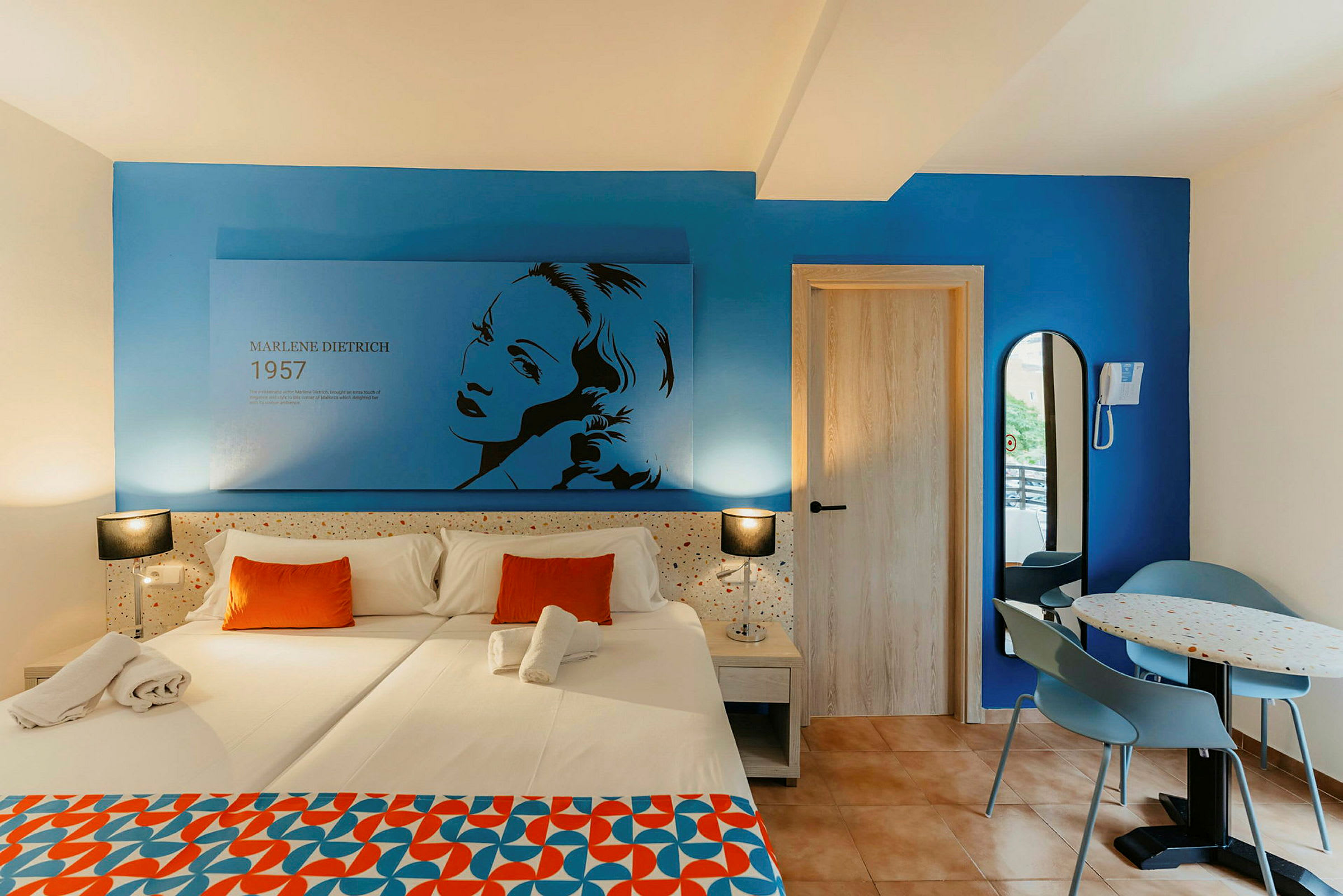 Et hotelværelse med en dobbeltseng med hvidt sengelinned og orange puder. Væggen er blå og har en illustration af Marlene Dietrich med tekst. Til højre er der en dør og en lille spiseplads med et bord og to stole. (AI-genereret tekst)