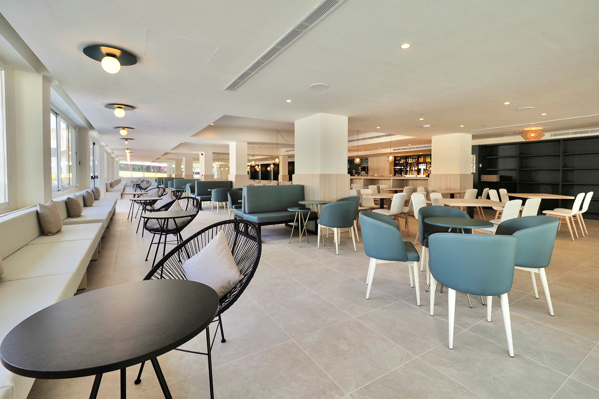 Billede af en moderne og rummelig indendørs lounge eller restaurant med borde og stole samt store vinduer, der lader dagslys strømme ind. (AI-genereret tekst)