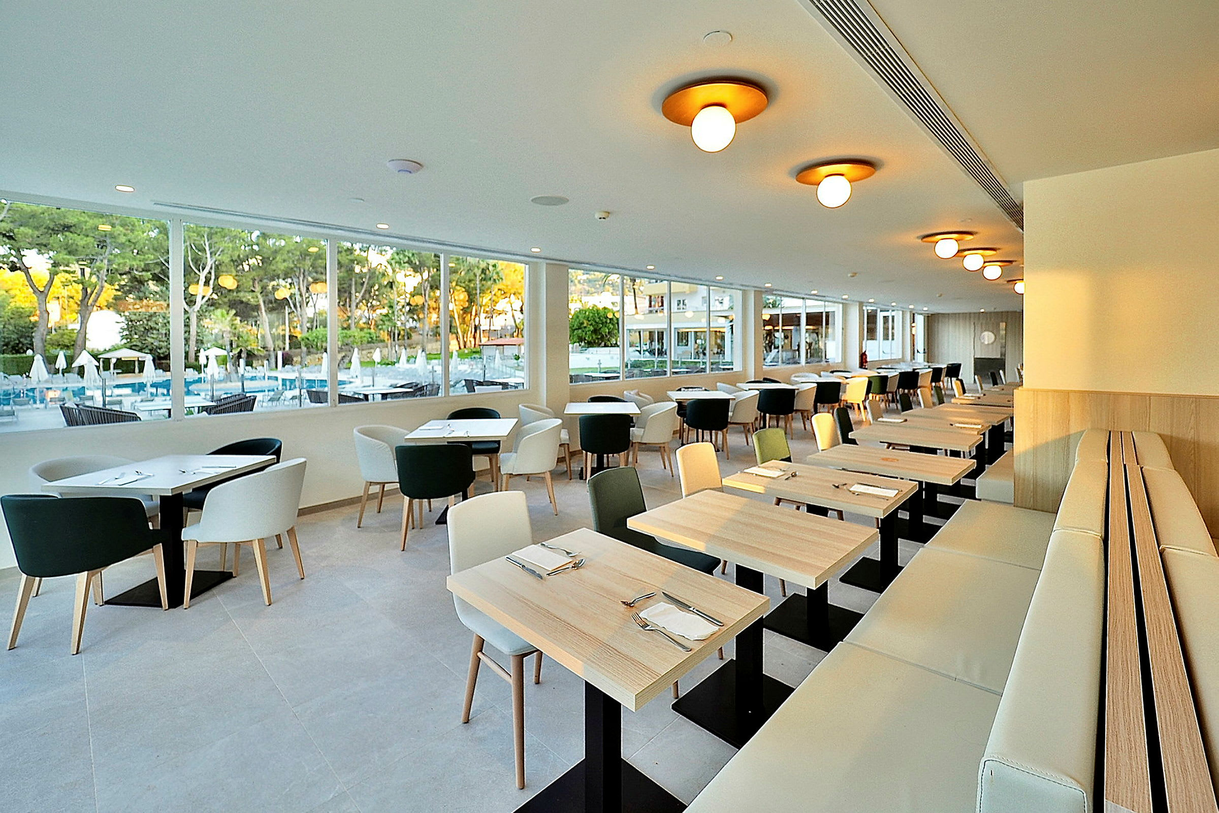 En moderne restaurant med vinduer, der vender ud mod en udendørs pool og grønne områder. Bordene og stolene er lyse og enkelt designet. (AI-genereret tekst)