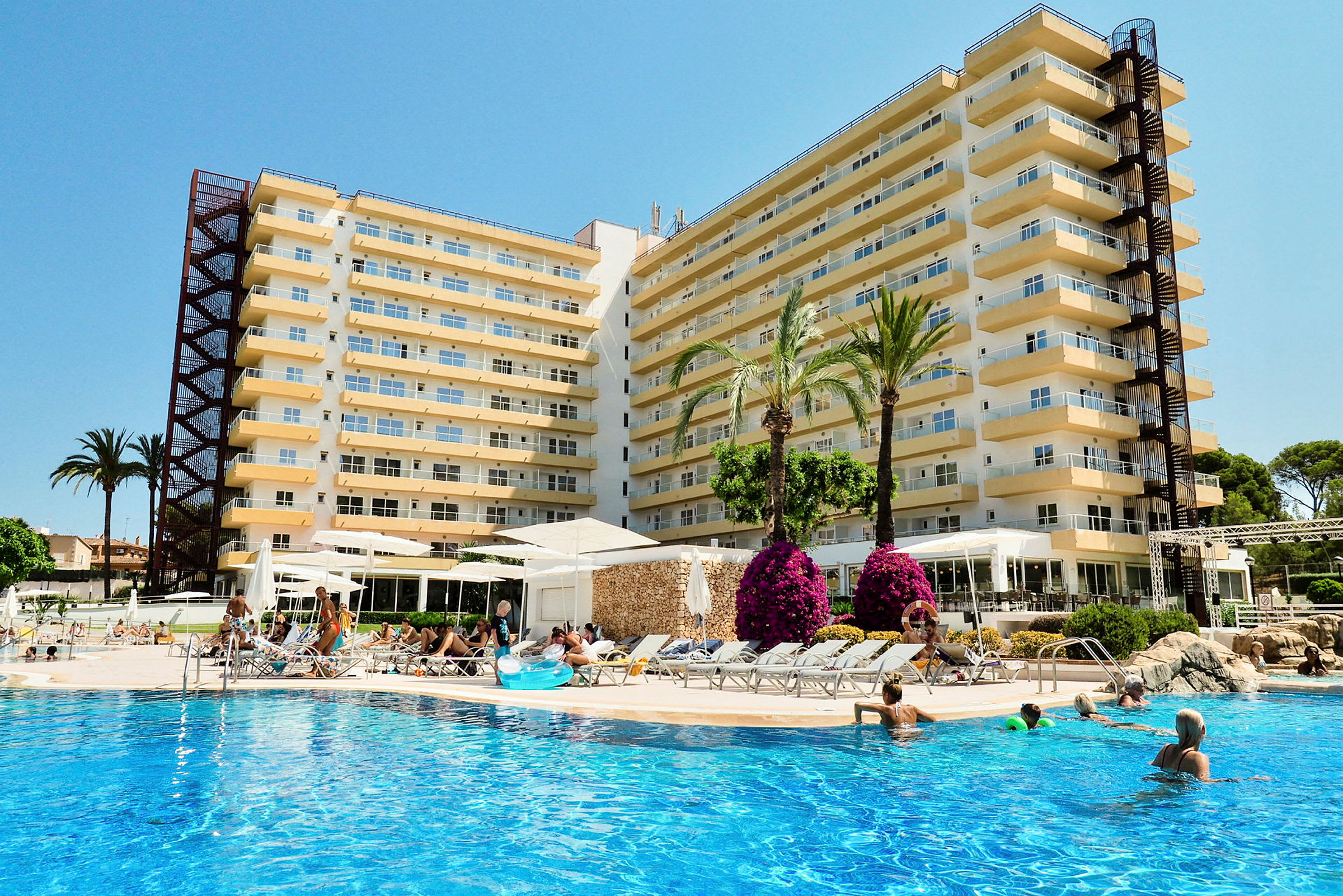 Billede af et hotel med flere etager, inklusive en udendørs pool, hvor folk svømmer og slapper af. Der er palmer og blomster omkring poolområdet. (AI-genereret tekst)