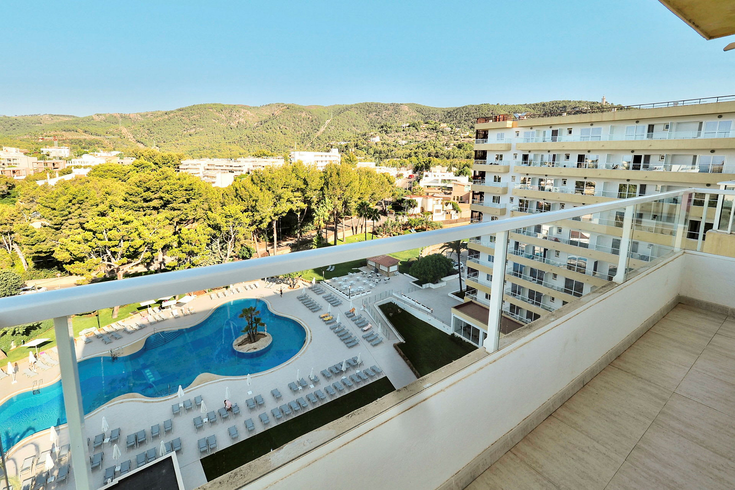 Udsigt fra en balkon over et hotelområde. Nederst ses en pool med liggestole og parasoller omkring. Boligbygninger og grønne bakker ses i baggrunden. (AI-genereret tekst)