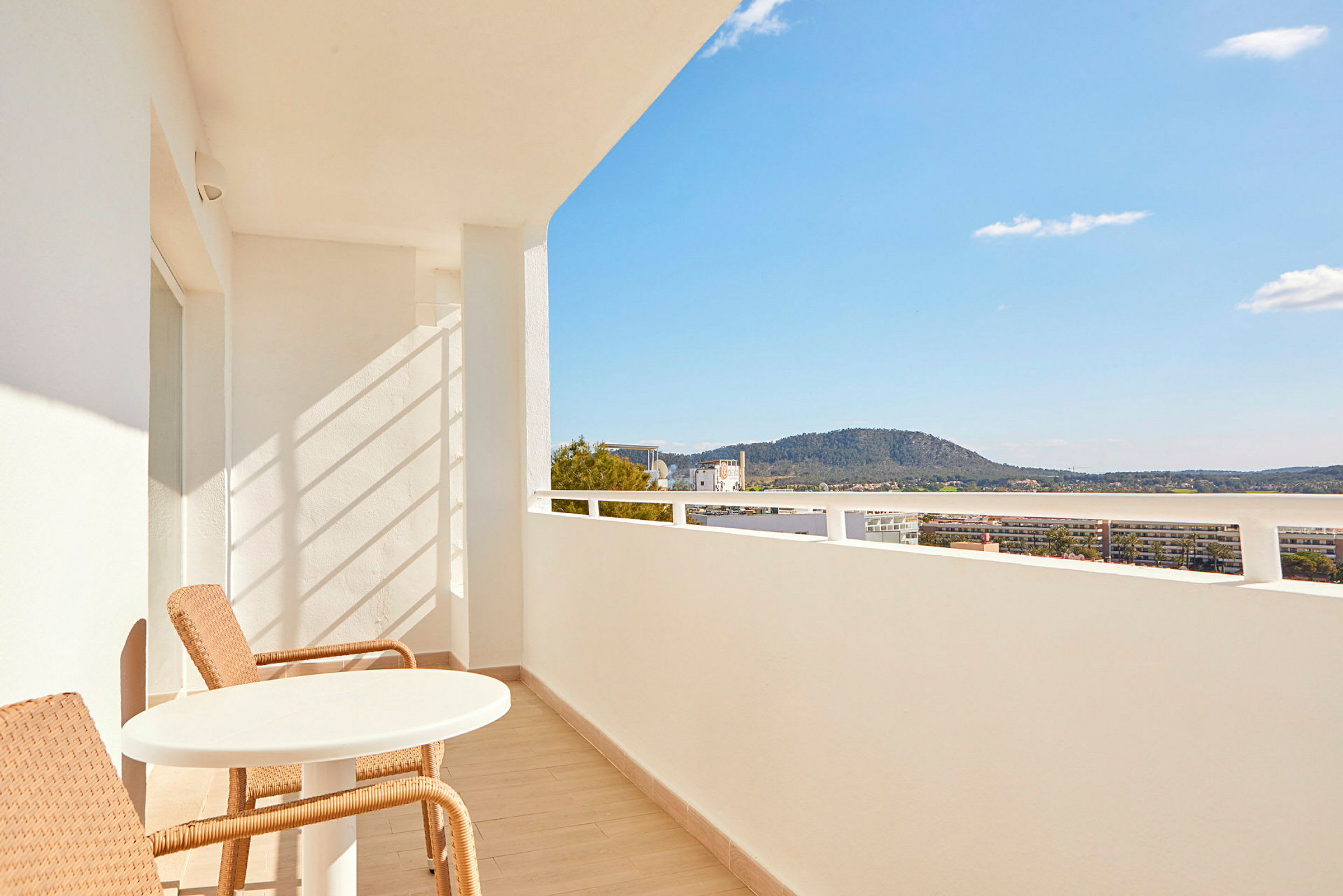 En balkon med hvide vægge, to flettede stole og et rundt bord, med udsigt over en bakke og den blå himmel. (AI-genereret tekst)