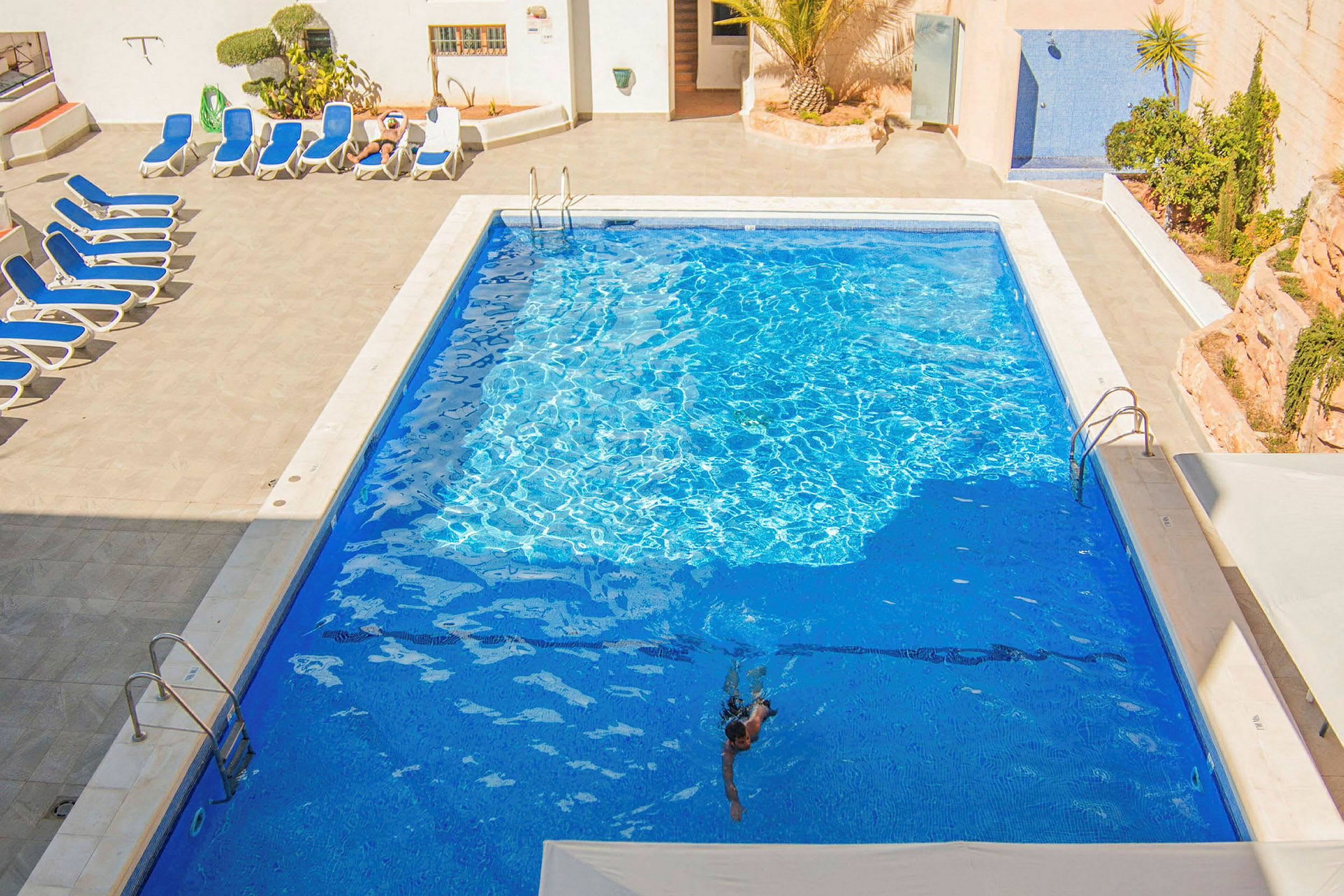 En udendørs swimmingpool med en nøgen mand, der svømmer, samt liggestole og planter ved poolkanten. (AI-genereret tekst)
