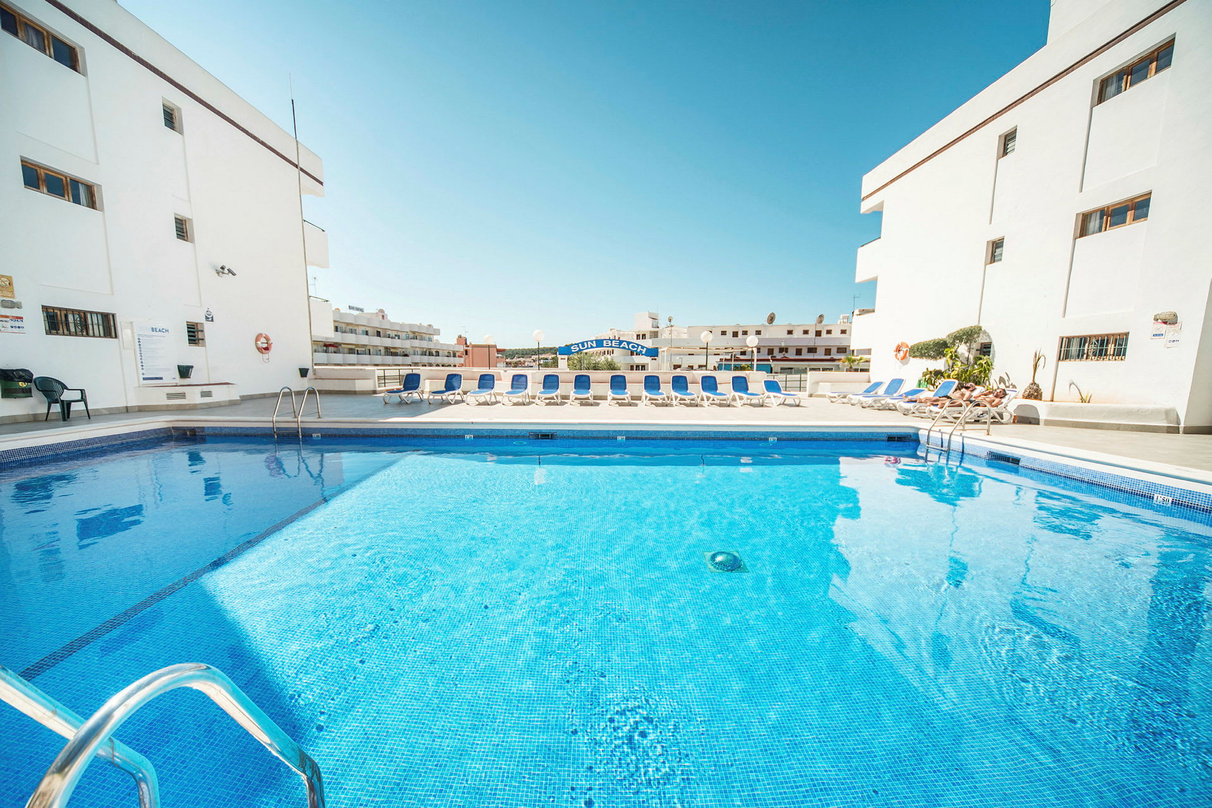 Billede af en swimmingpool omgivet af liggestole og hvide bygninger. (AI-genereret tekst)