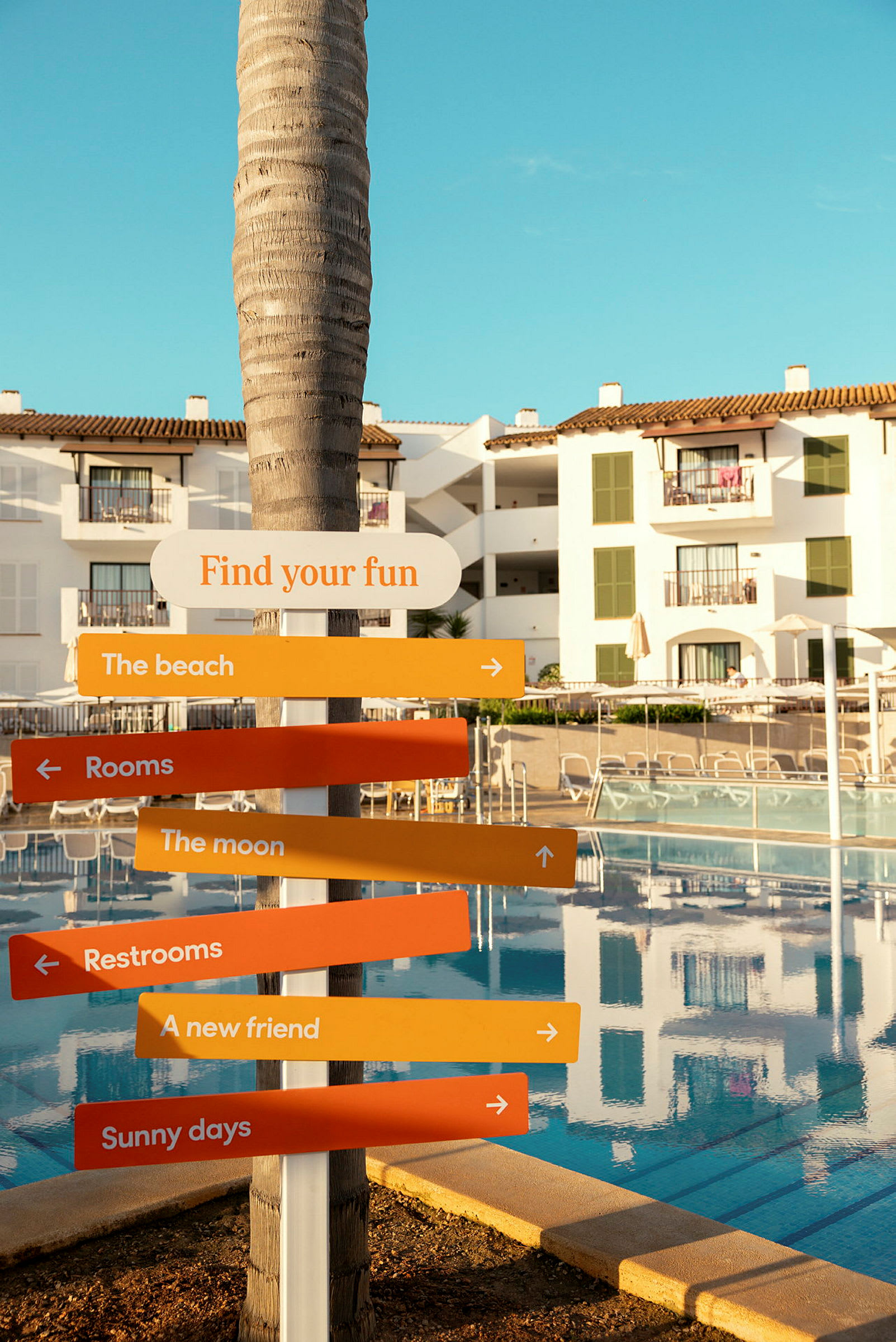 Et skilt med teksten 'Find your fun' og flere farvede pile på en resort med pool og hvide bygninger. (AI-genereret tekst)