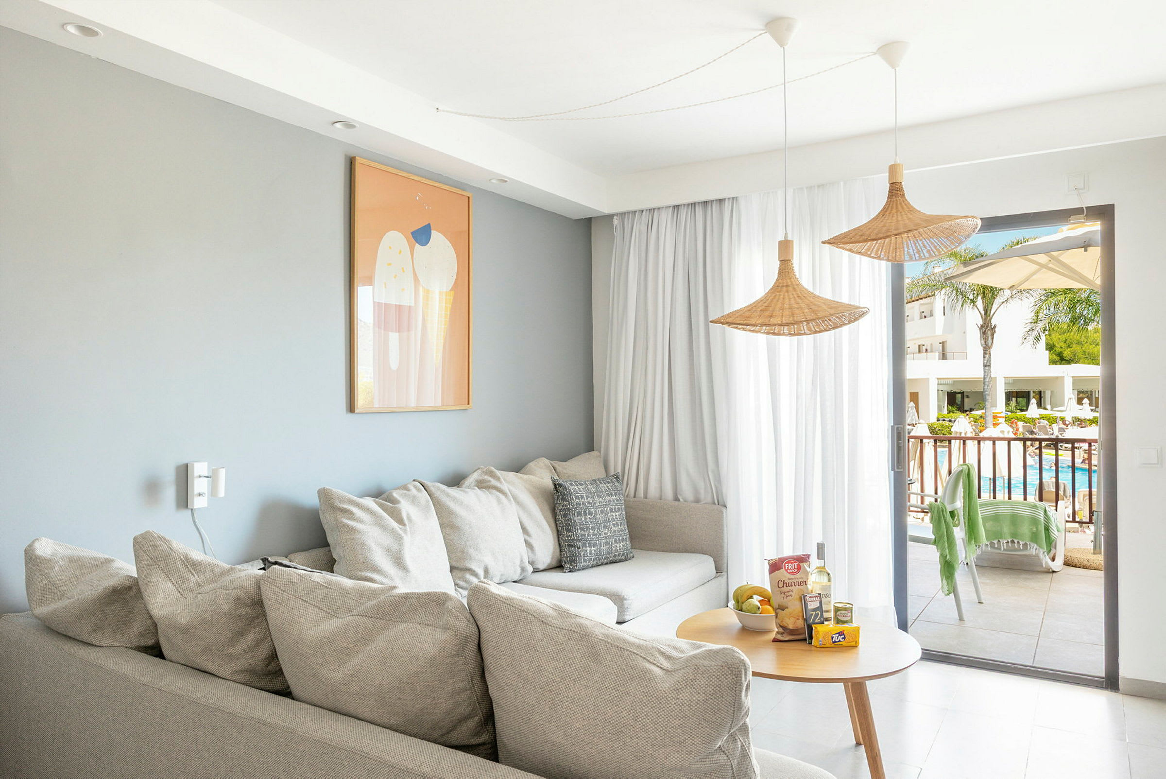 Stue med sofa, et lille bord og en åbning mod en balkon med udsigt over en pool. (AI-genereret tekst)