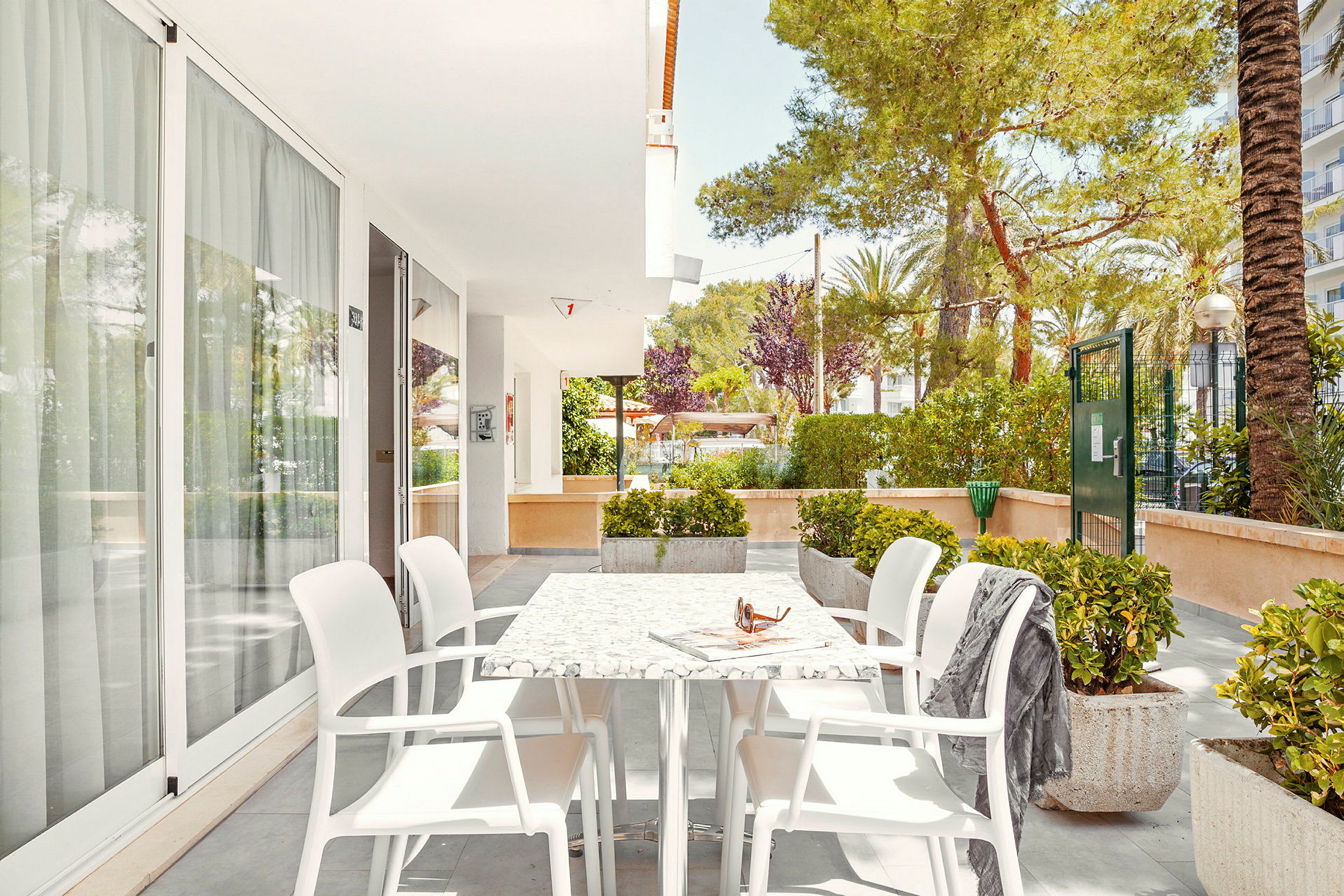 Udendørsområde med bord og stole på en flisebelagt terrasse, omringet af grønne omgivelser. (AI-genereret tekst)