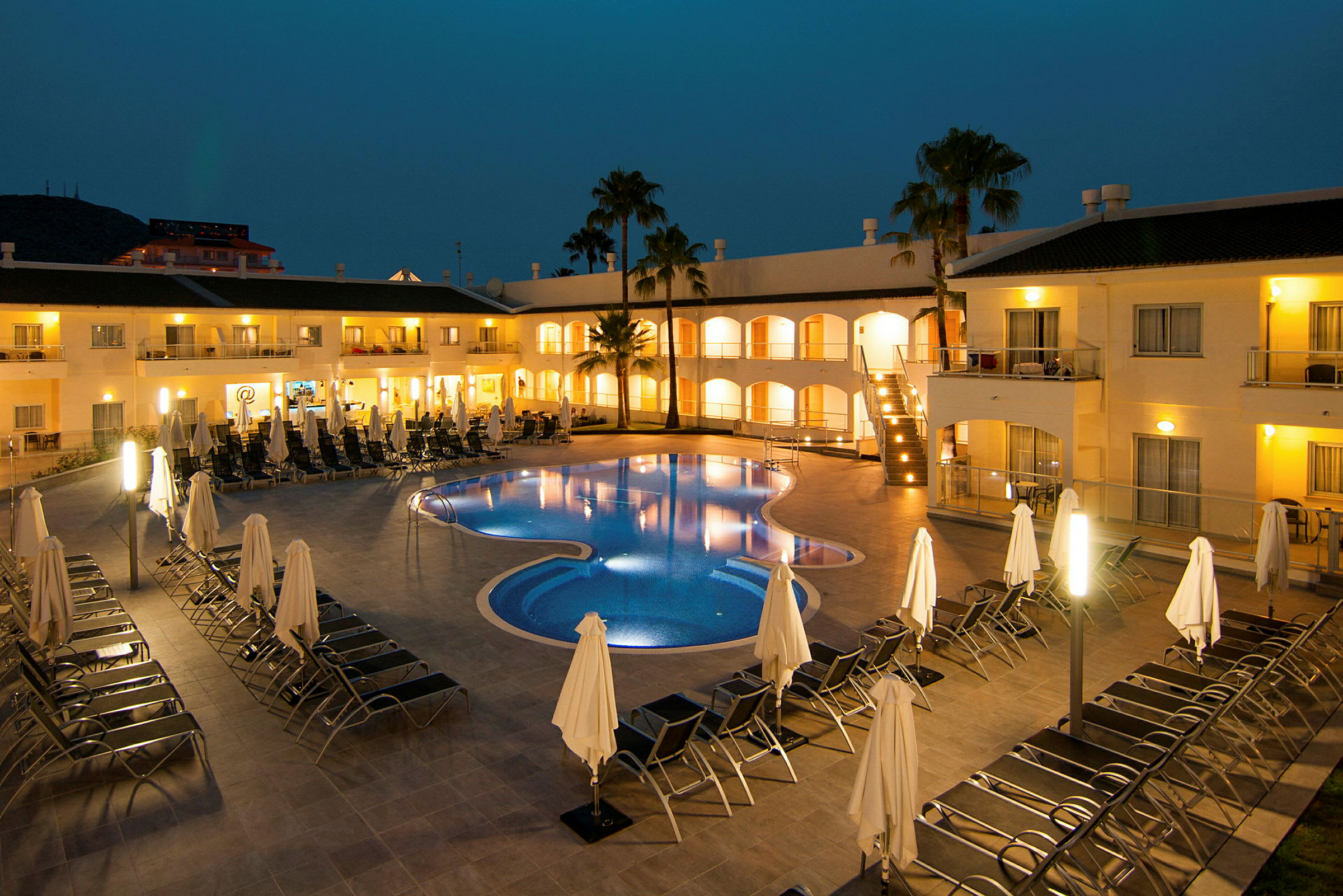 Hotel med oplyst udendørs pool omgivet af liggestole og parasoller om aftenen. (AI-genereret tekst)