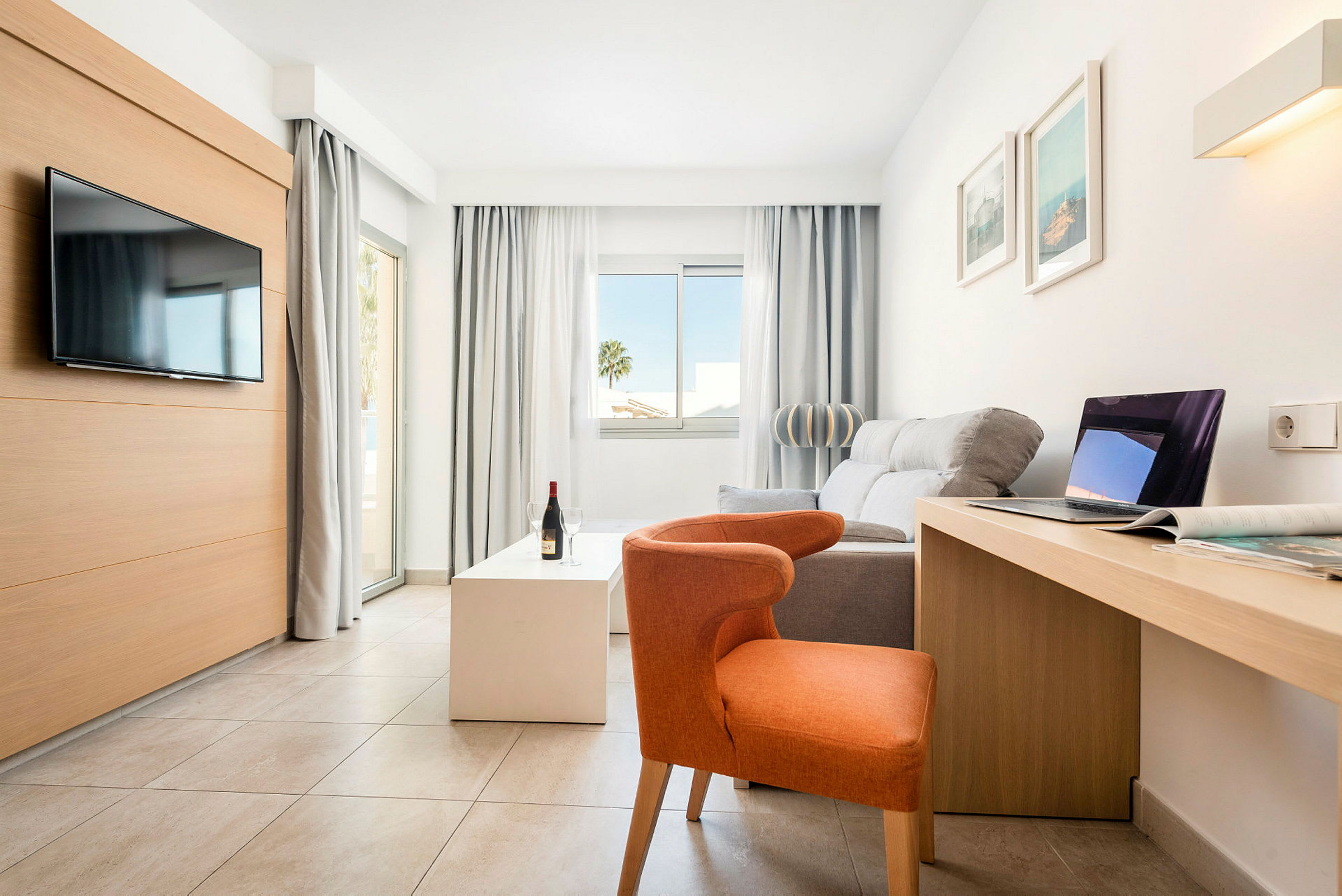 Lyst, moderne hotelværelse med sofa, stol, TV, skrivebord og store vinduer. (AI-genereret tekst)