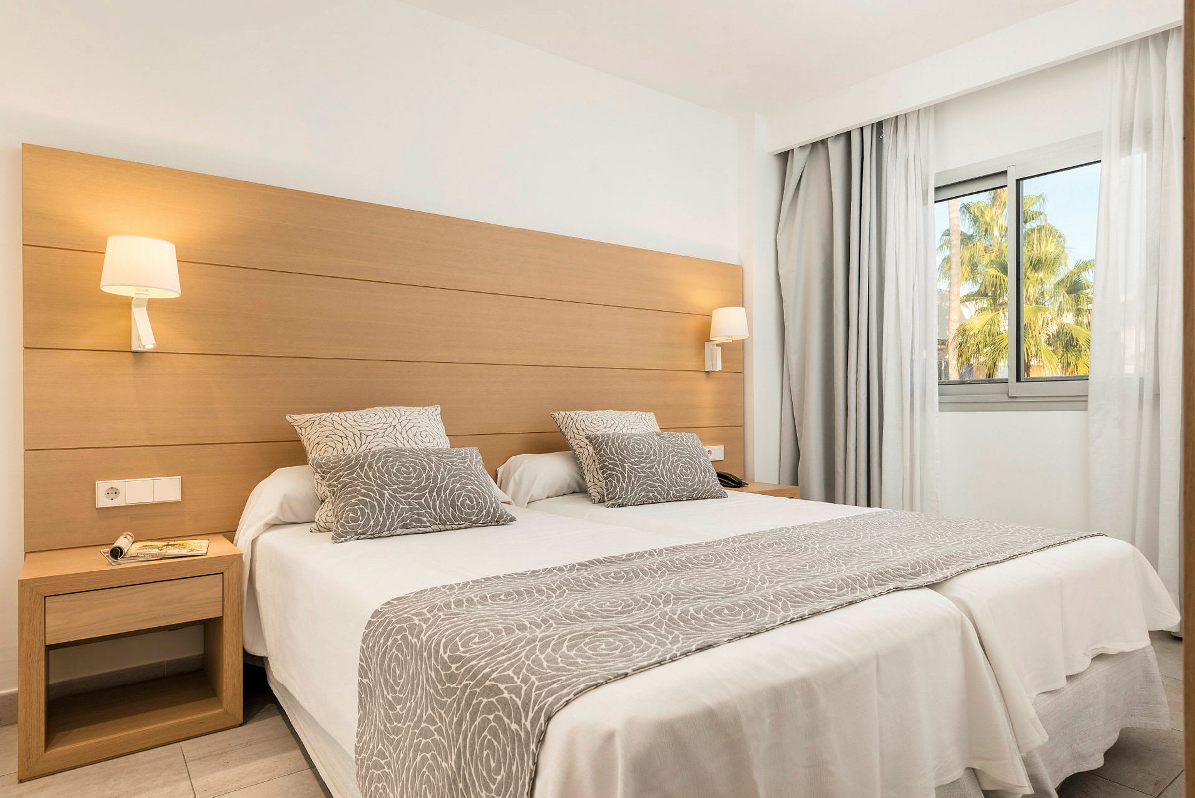 Et hotelværelse med to enkeltsenge sat sammen, dekoreret med gråhvide tekstiler med mønster, og et stort vindue med udsigt til grønne områder. (AI-genereret tekst)